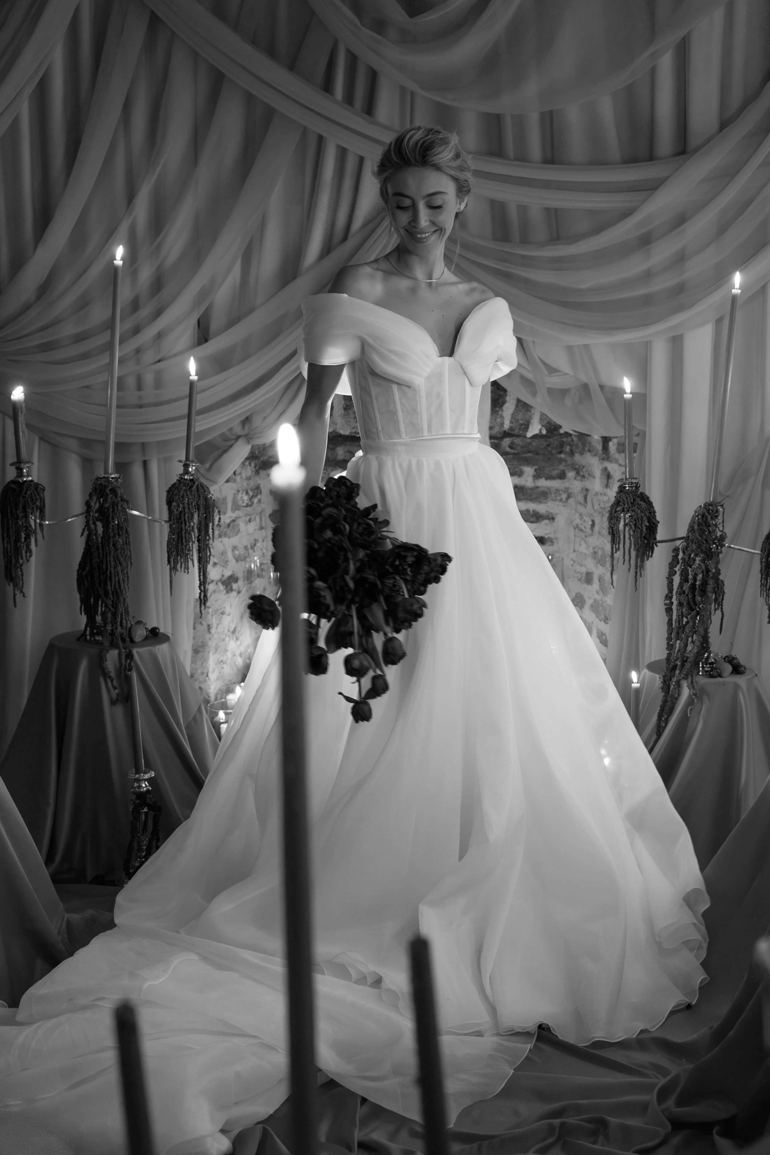 Black and white photo of a woman in a wedding dress holding a bouquet of dark roses, standing in front of a draped fabric backdrop with candles and tables with plants.