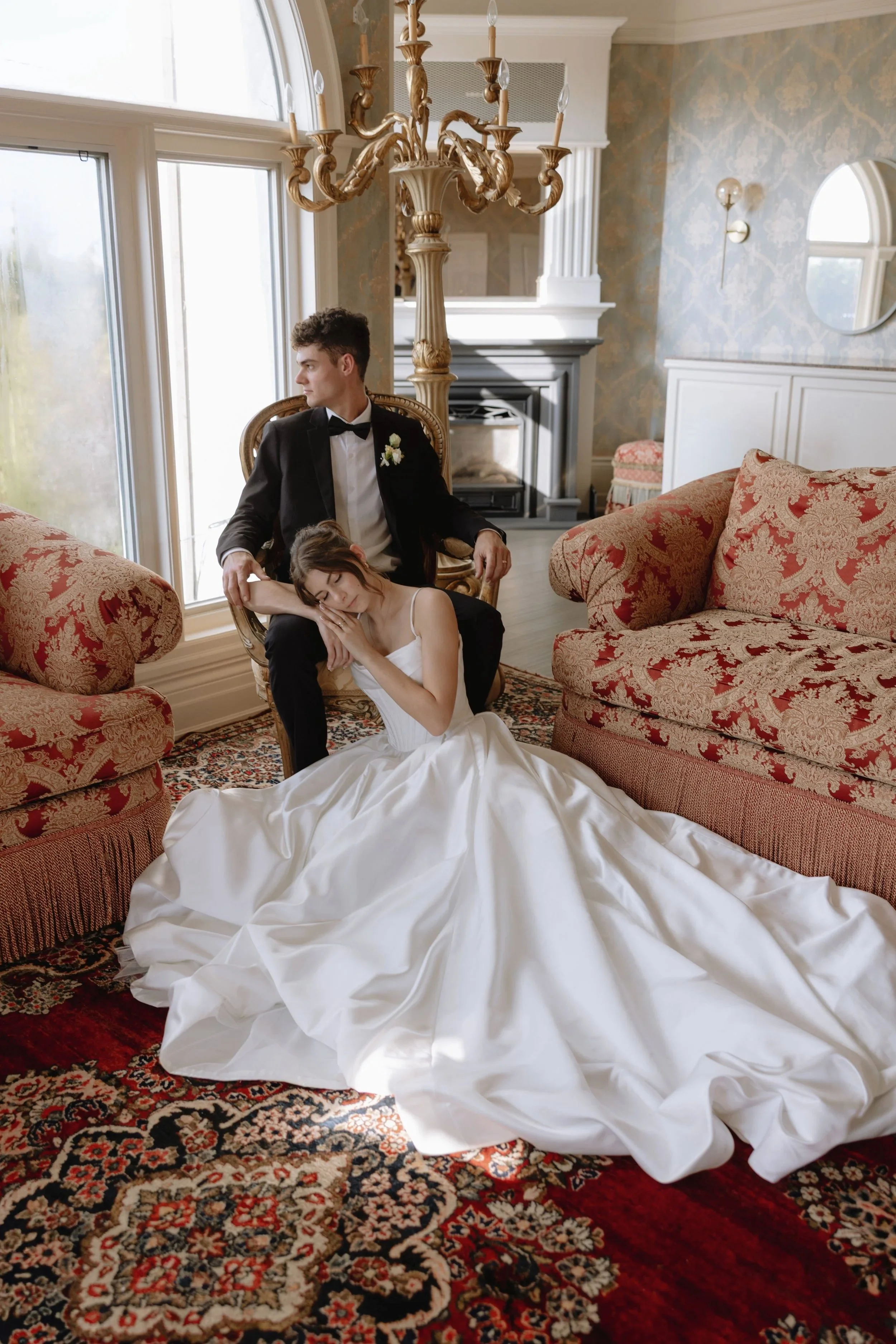 A bride in a white wedding gown sleeping with her head on the groom's lap, who is sitting on a gold-colored chair in a decorated living room. The groom is dressed in a black tuxedo with a white shirt and bow tie at an estate venue Niagara Ontario.