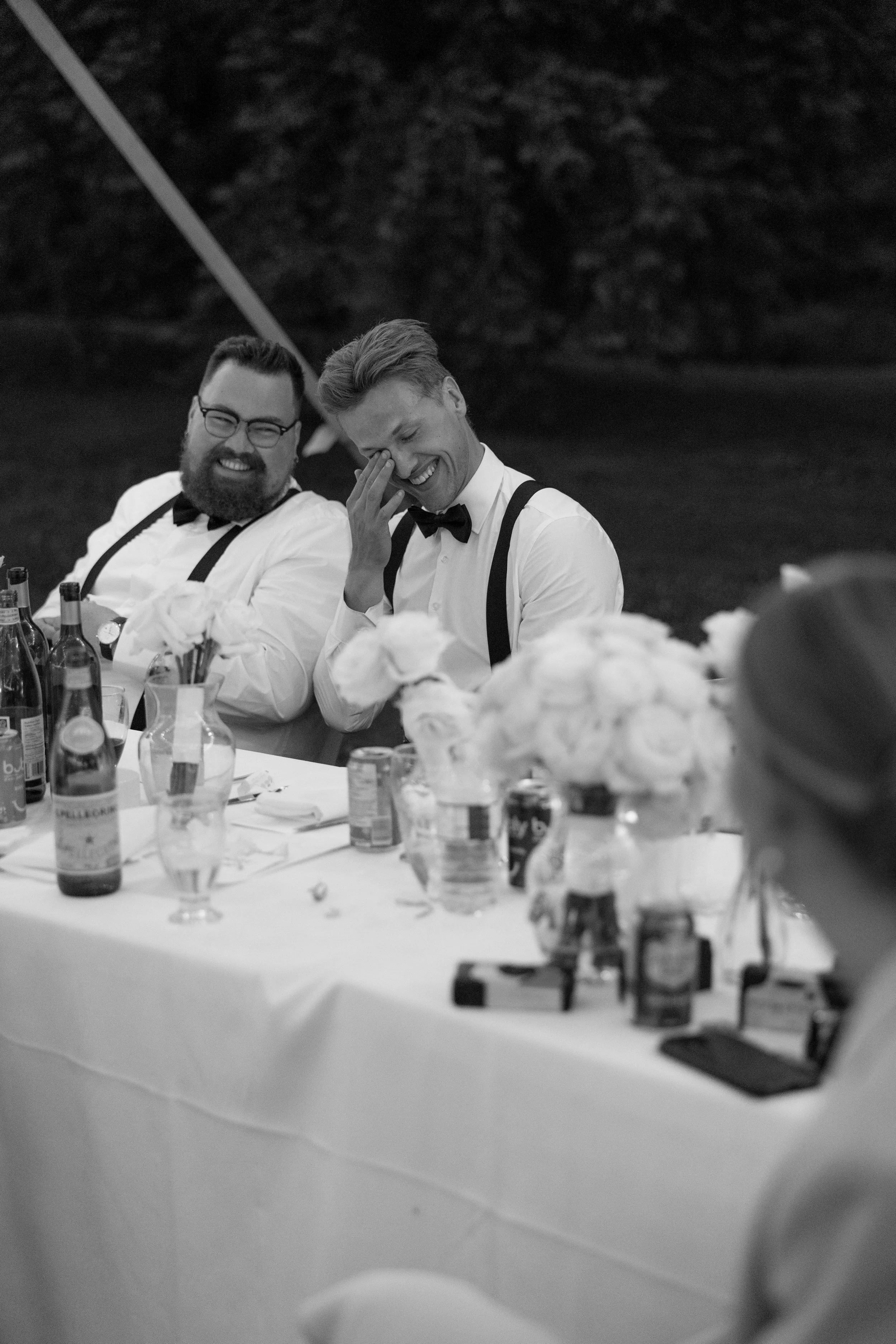 Two men dressed in white shirts with suspenders and bow ties are sitting at a long table outdoors. They are smiling and laughing, with one man touching his face. The table is decorated with flowers and drinks at a backyard wedding in the GTA Ontario.