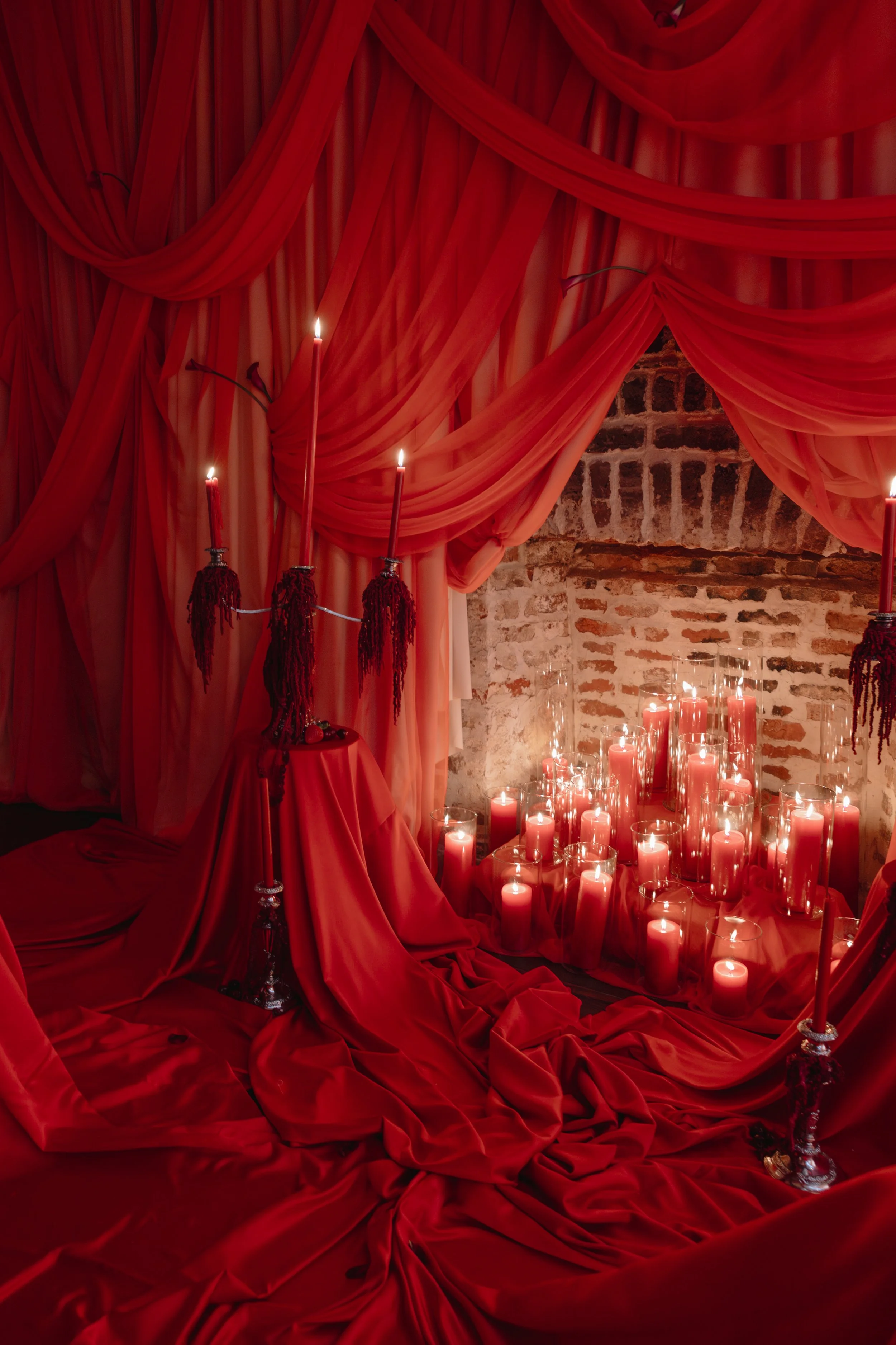 A romantic scene with red draped fabric, tall red candles, and numerous candles in glass containers on the floor, all illuminated by candlelight, against an exposed brick wall for a wedding reception in Charleston, South Carolina.