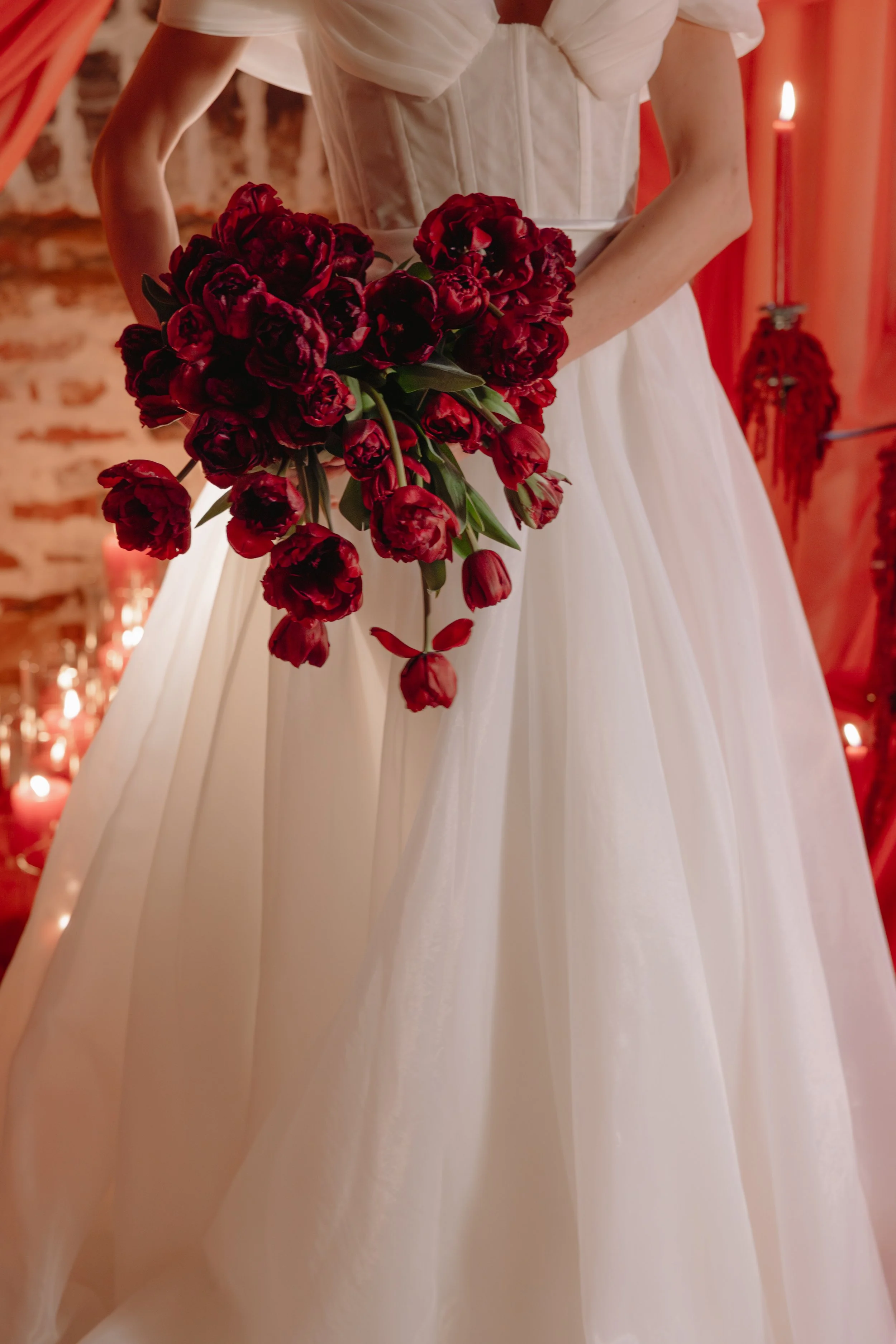 A bride in a white wedding gown holding a bouquet of dark red tulips, standing in a decorated indoor setting with candles and candles in the background  for a wedding reception in Charleston, South Carolina.