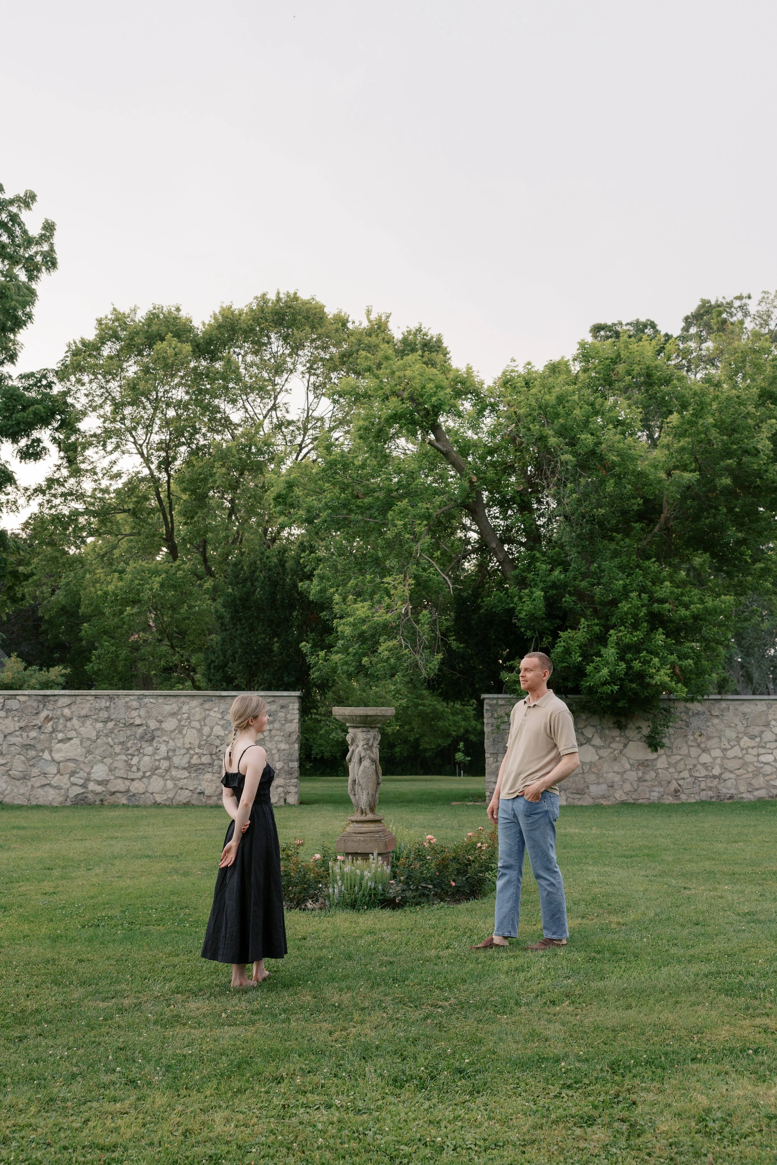 A man and woman dancing on a stone path in a garden with trimmed bushes and tall trees, with a large two-story house with a porch in the background. An engagement session in Hamilton Ontario. 