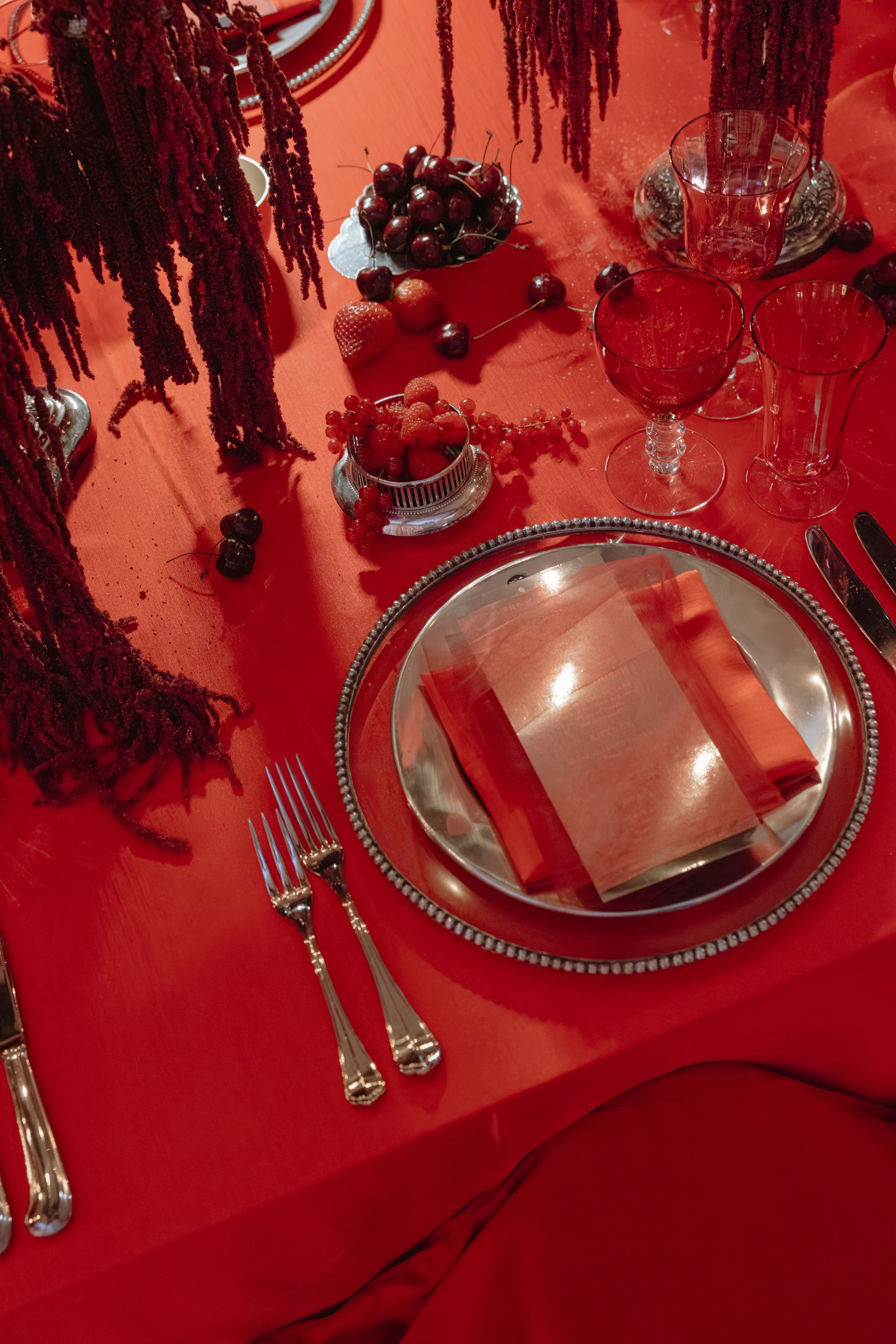 Set dining table with red tablecloth, silver charger plate, red napkin, and silver cutlery. Glassware includes a red tinted wine glass and a clear glass. Centerpieces feature red and black berries, strawberries, and cherries. Decor includes hanging r