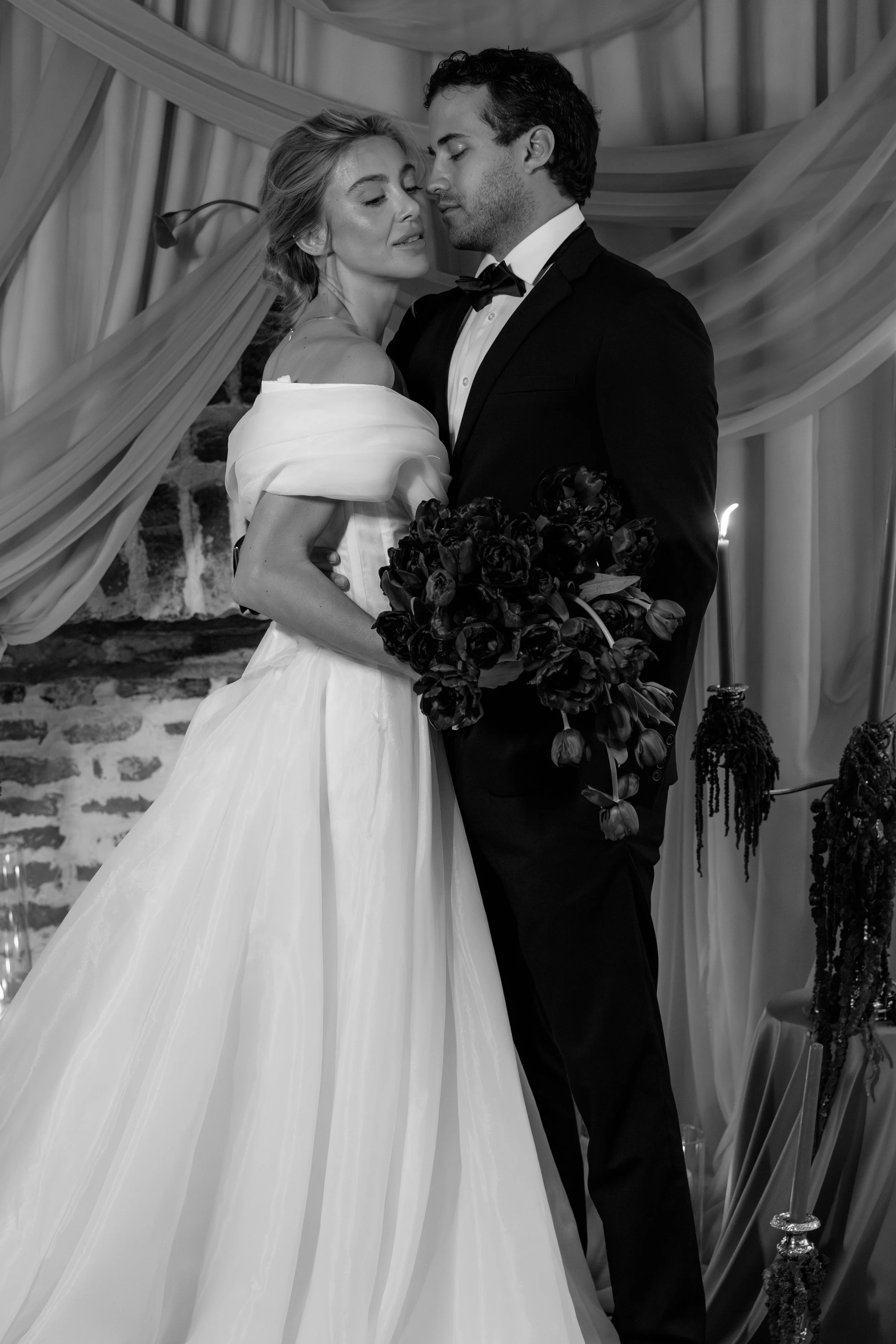 Black and white photo of a bride in a wedding gown and a groom in a tuxedo, holding a bouquet of flowers, standing close together and touching foreheads at a wedding reception in Charleston, South Carolina.