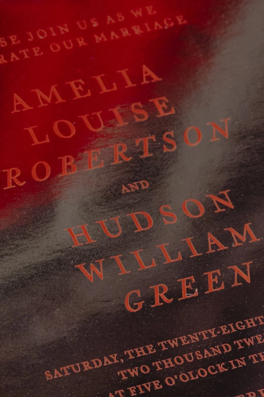 Close-up of a wedding invitation or event poster with red and black background, featuring names Amelia Louise Robertson and Hudson William Green in bold red text, and date details at the bottom for a wedding reception in Charleston, South Carolina.