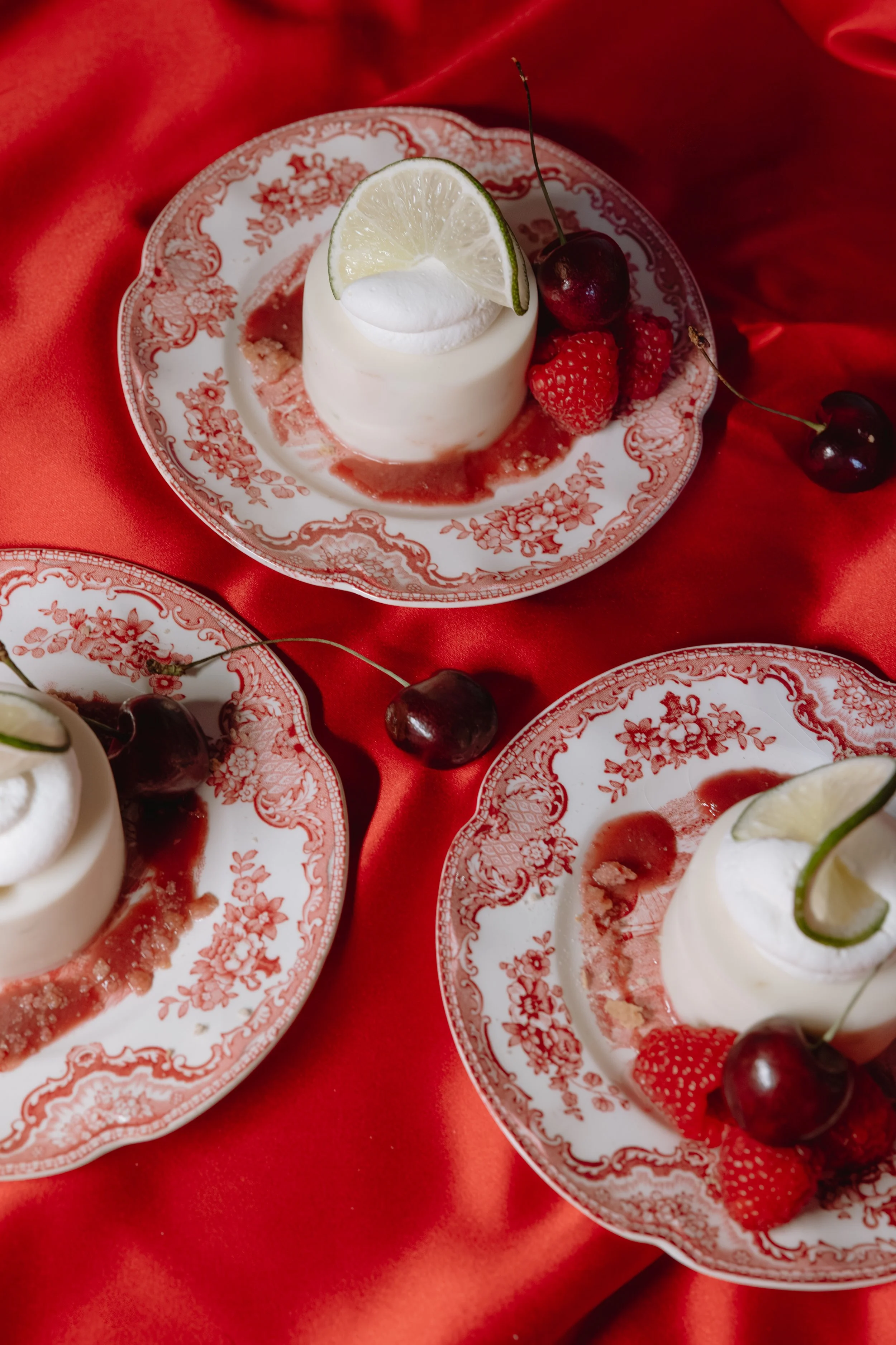 Three plates of panna cotta topped with a lime wedge and fresh cherries and raspberries on a red satin cloth.