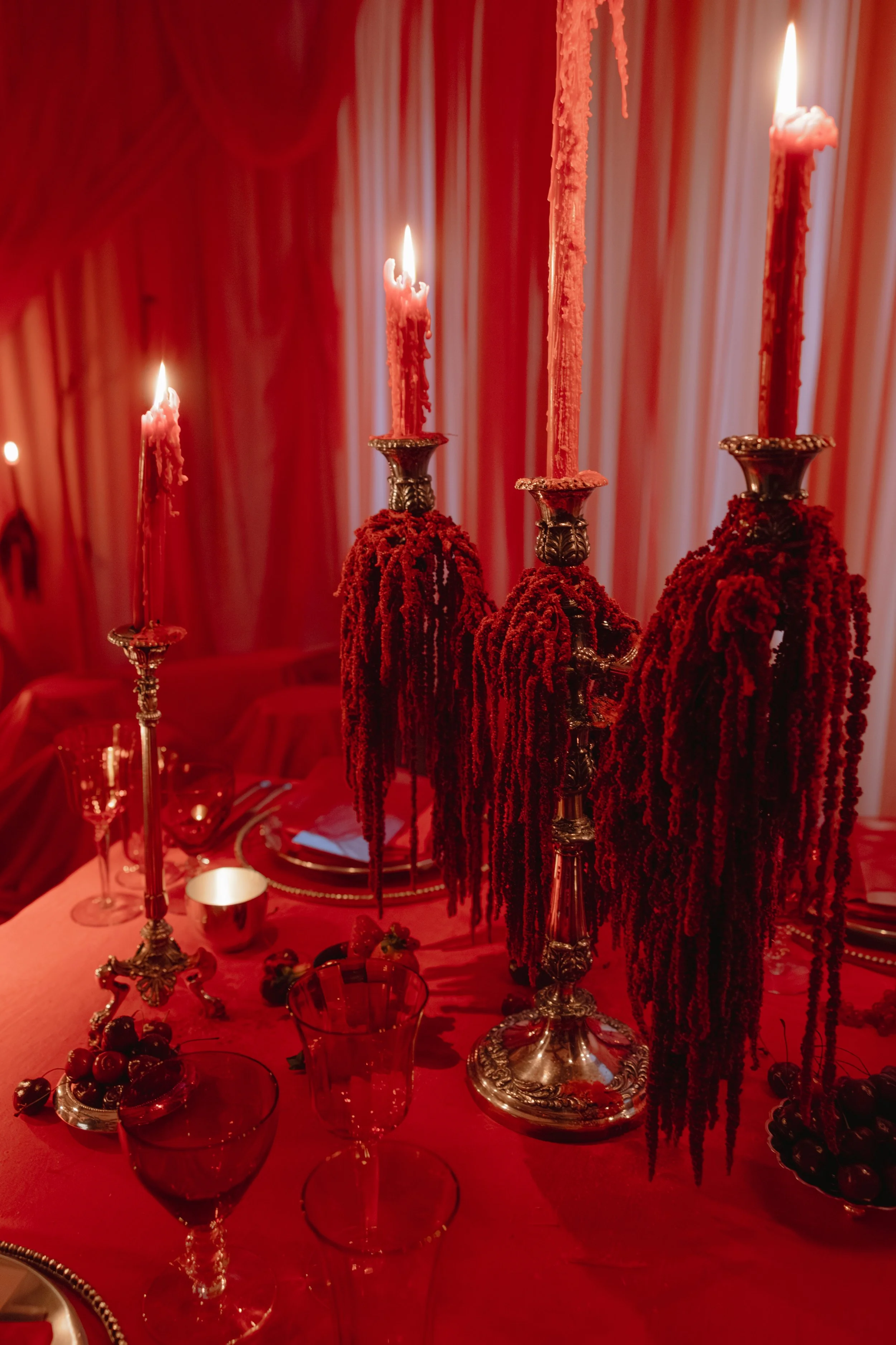 Elegant red-themed table setting with three ornate silver candle holders and tall pink candles, surrounded by red grapes, glassware, and red curtains in the background  for a wedding reception in Charleston, South Carolina.