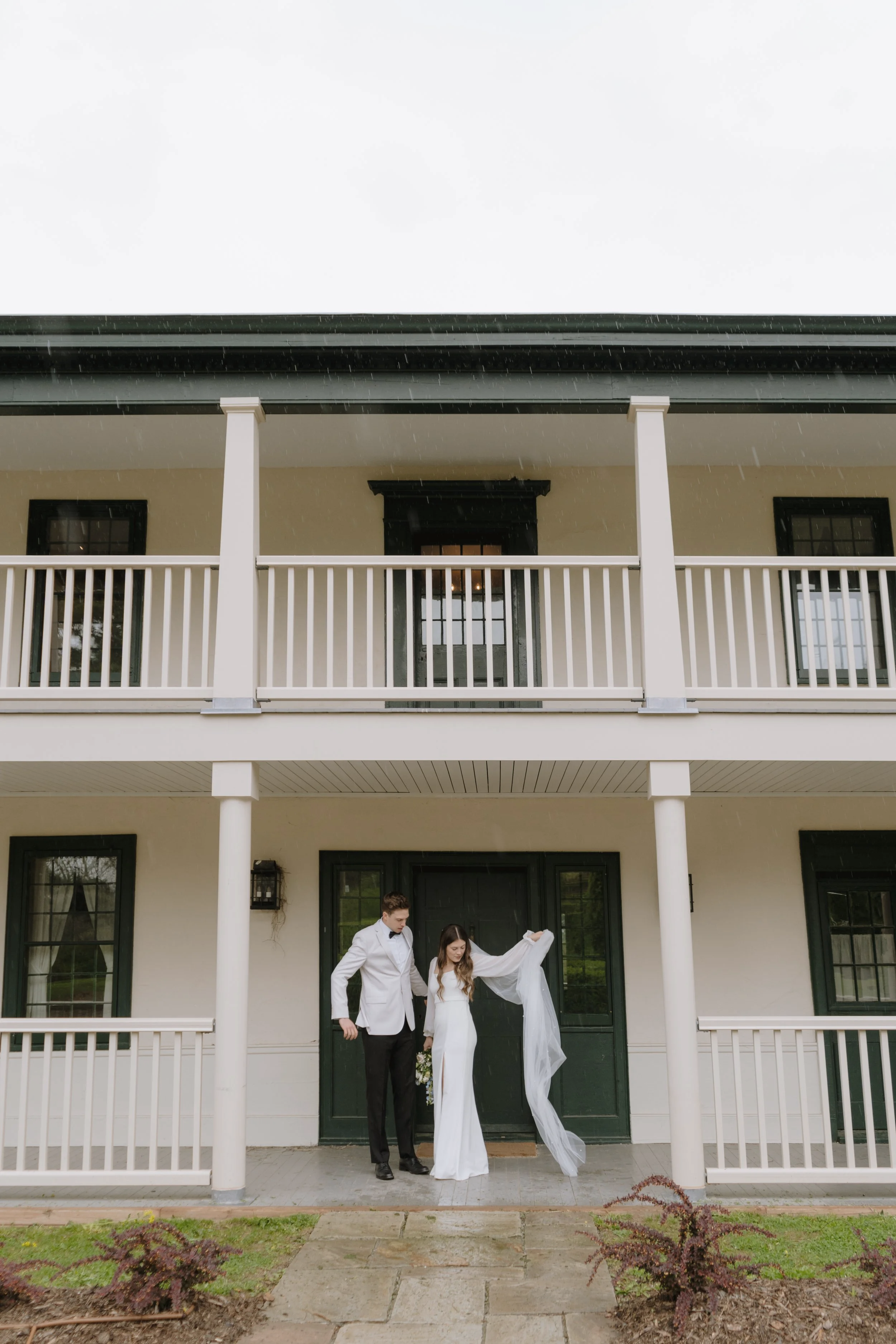 A bride and groom standing on the porch outside a two-story house in the rain, with the groom in a white tuxedo and the bride in a white wedding dress holding up a veil for a wedding in Hamilton Ontario.