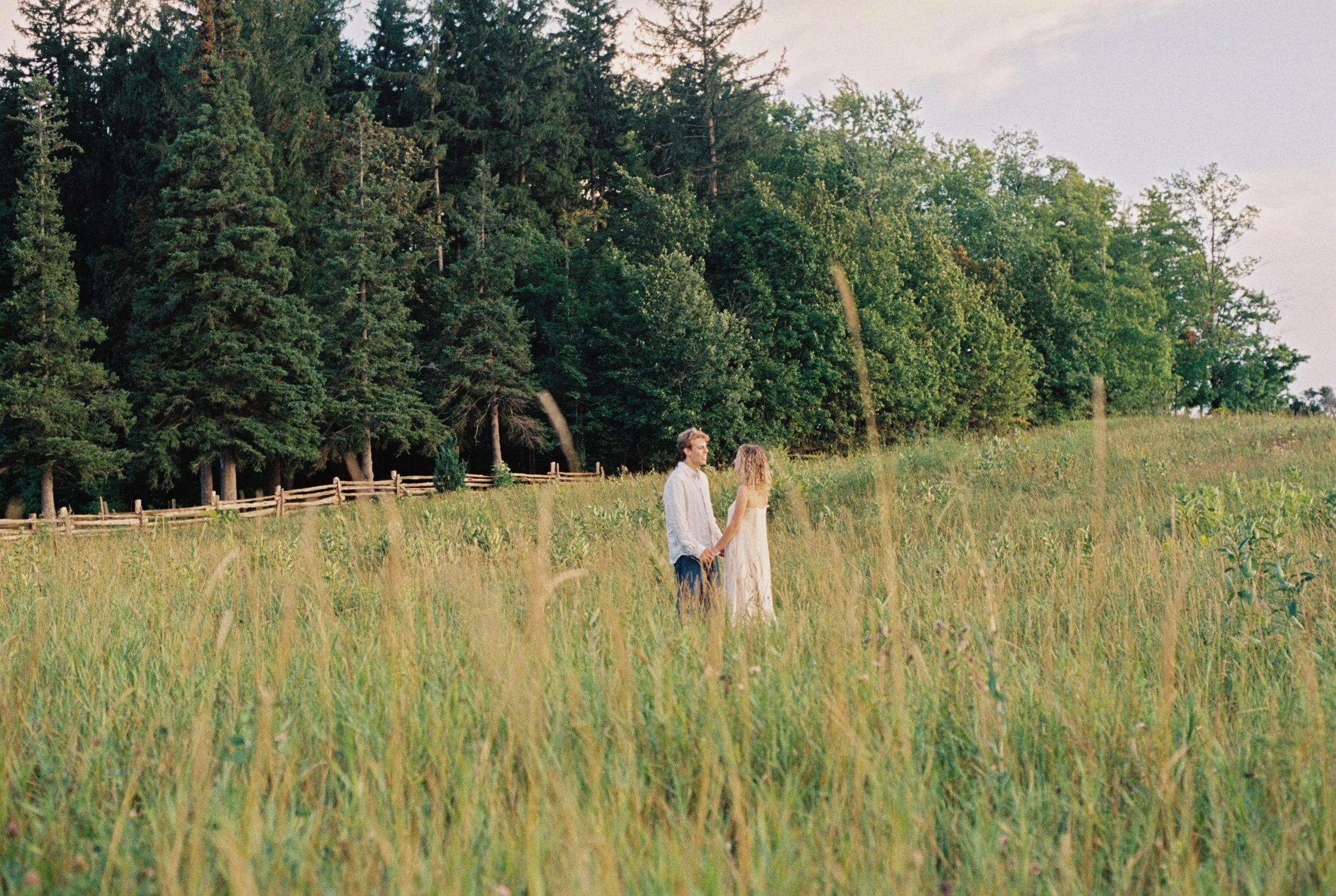A man and woman dancing on a stone path in a garden with trimmed bushes and tall trees, with a large two-story house with a porch in the background. An engagement session in Hamilton Ontario. 