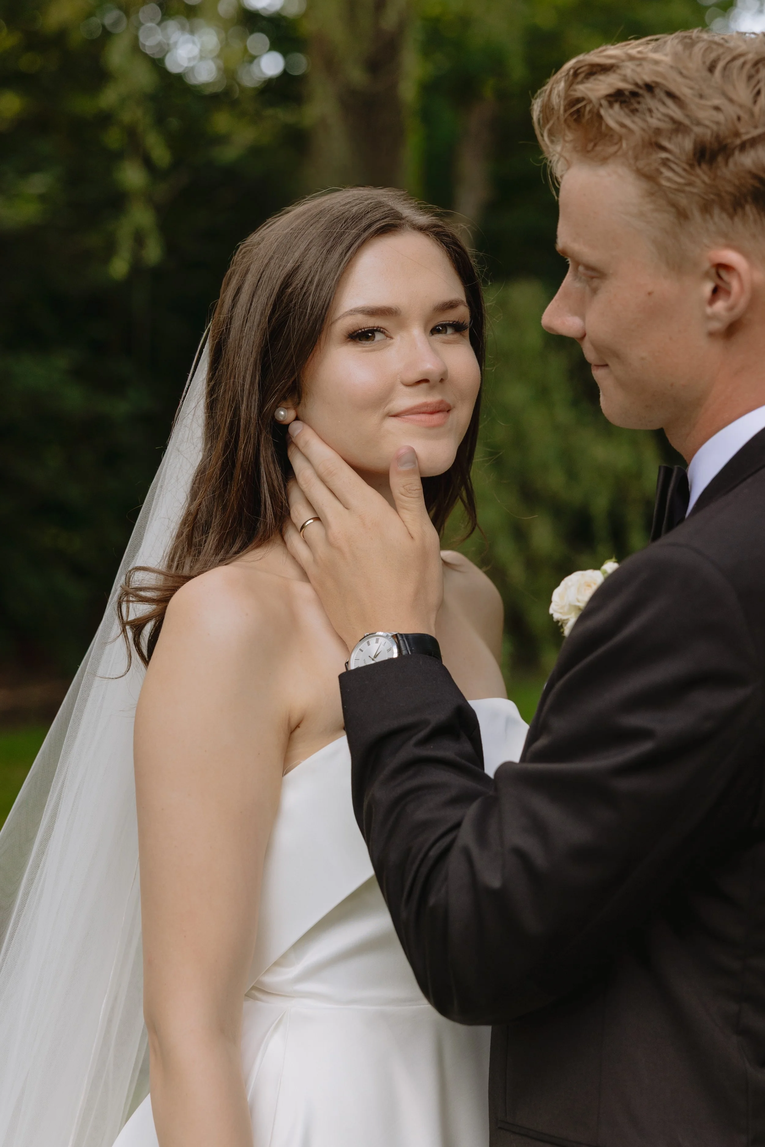 A groom gently touching his bride's chin outdoors, both dressed in wedding attire, with trees and greenery in the background for a backyard wedding in the GTA.