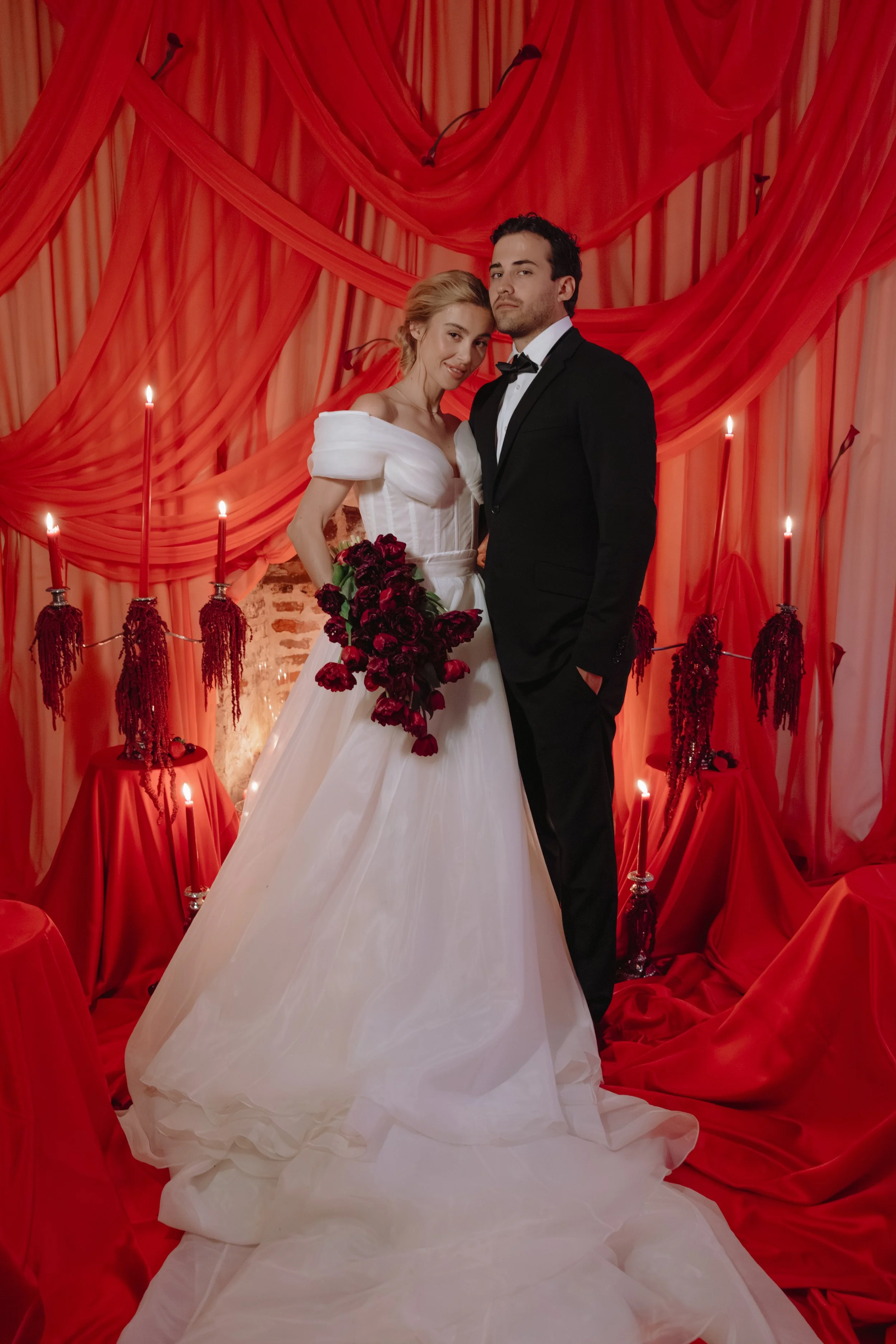 A bride and groom standing together in front of a red draped backdrop with candles and red decorations. The bride is wearing a white wedding dress with an off-shoulder design and holding a bouquet of dark red flowers. The groom is dressed in a black 
