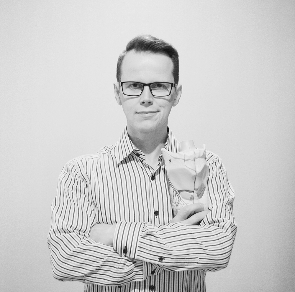 A man with glasses and a striped shirt holding a model of a human heart in front of a plain background.