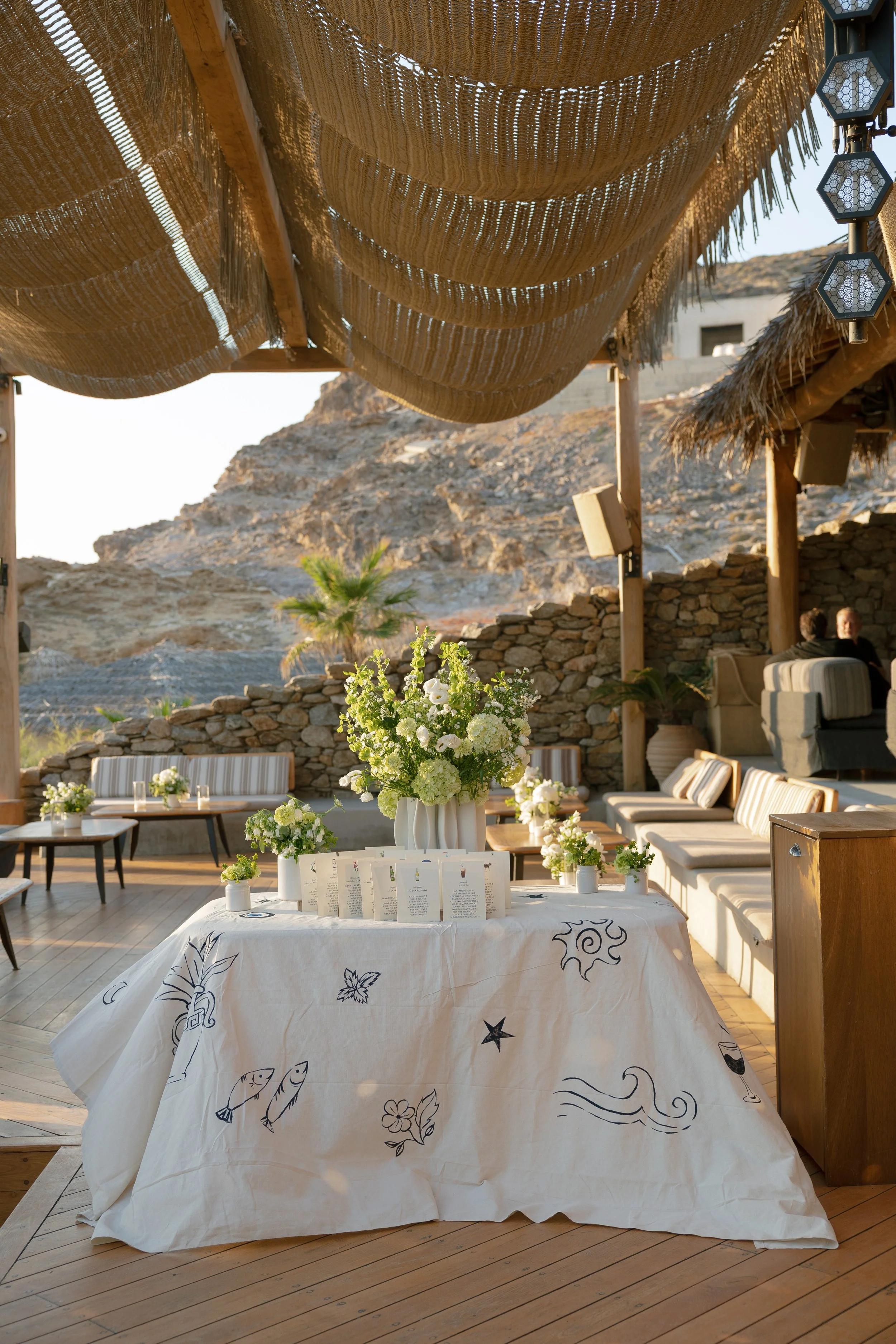 A hand-painted tablecloth decorated with black drawings of fish, flowers, and swirls, set with custom Greek menus for a wedding in Mykonos