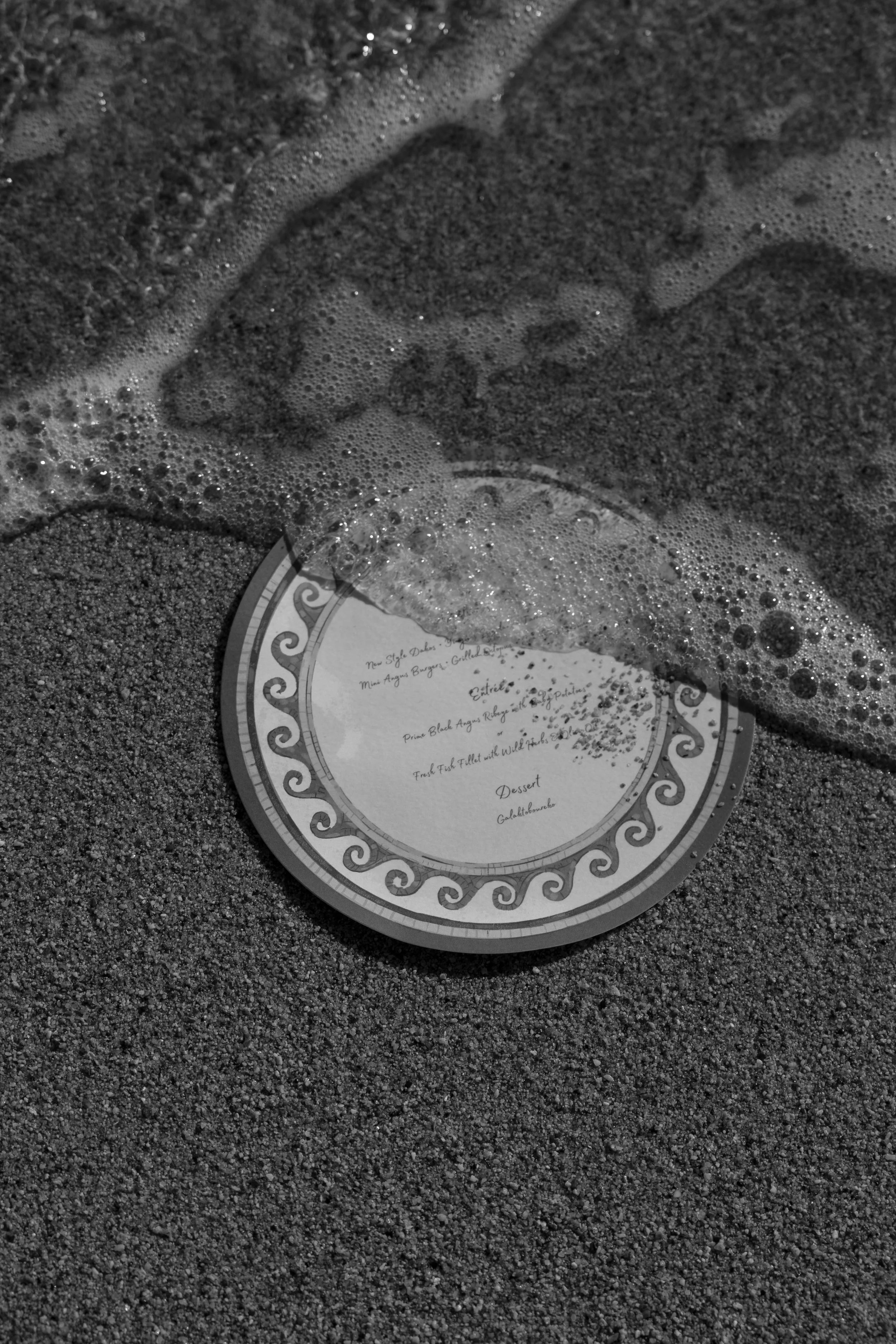 Custom circular menu with a decorative border partially submerged in sand on the beaches of Mykonos Greece