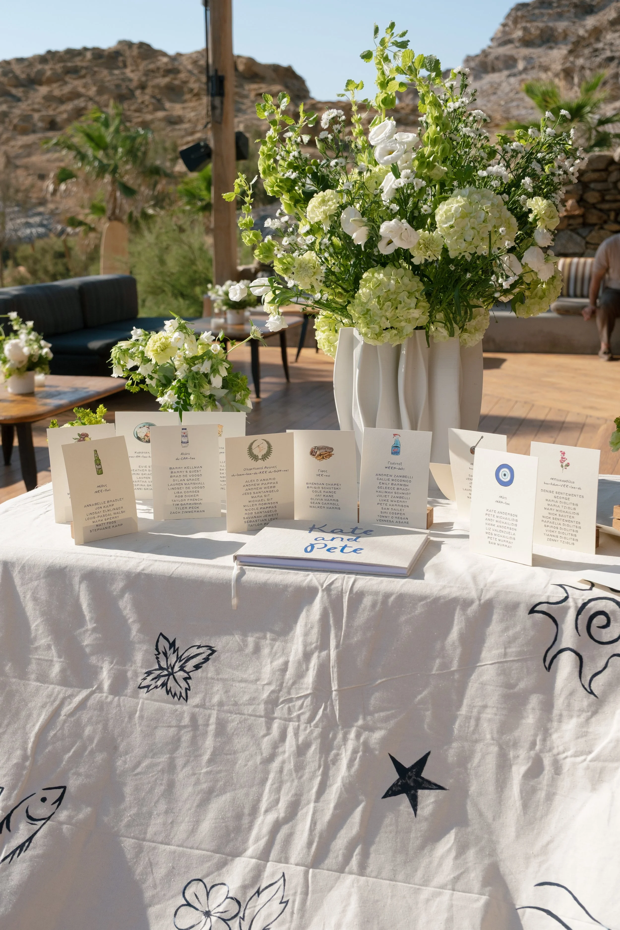 A table with a white cloth featuring black illustrations of fish, stars, and leaves, displaying multiple name cards and a large floral arrangement of white flowers in vases against an outdoor desert backdrop with rocks and mountains.