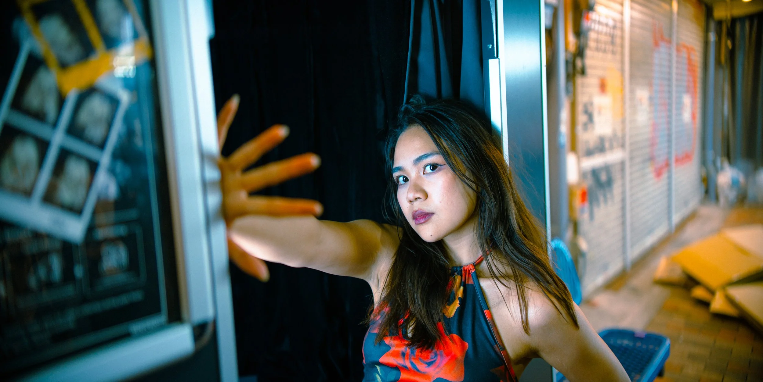 A woman with dark hair and a floral dress reaching out with her hand towards a vending machine in a store or market.