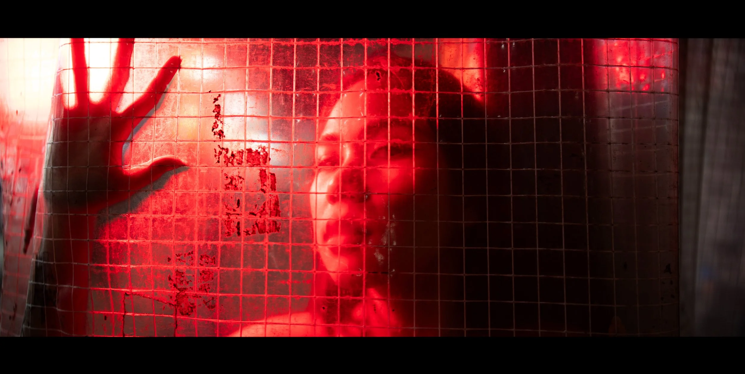 Person with short dark hair pressing their face and hand against a red-lit grid-covered glass surface, looking through it.