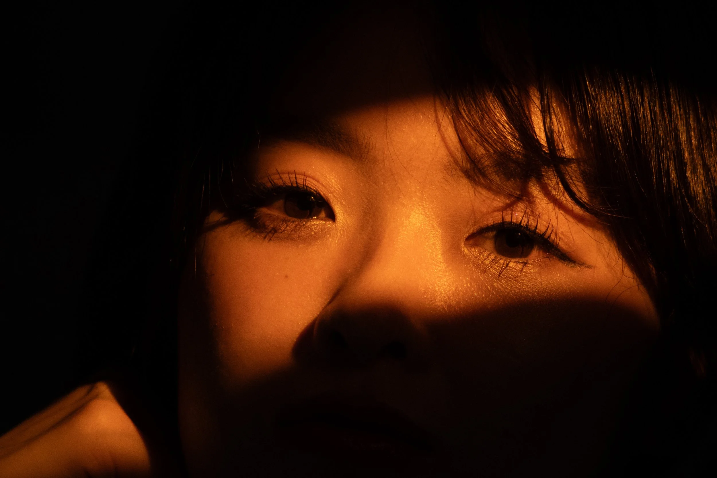 Close-up of a person's face illuminated by warm, golden light, with their eyes partially visible and shadows covering parts of the face.