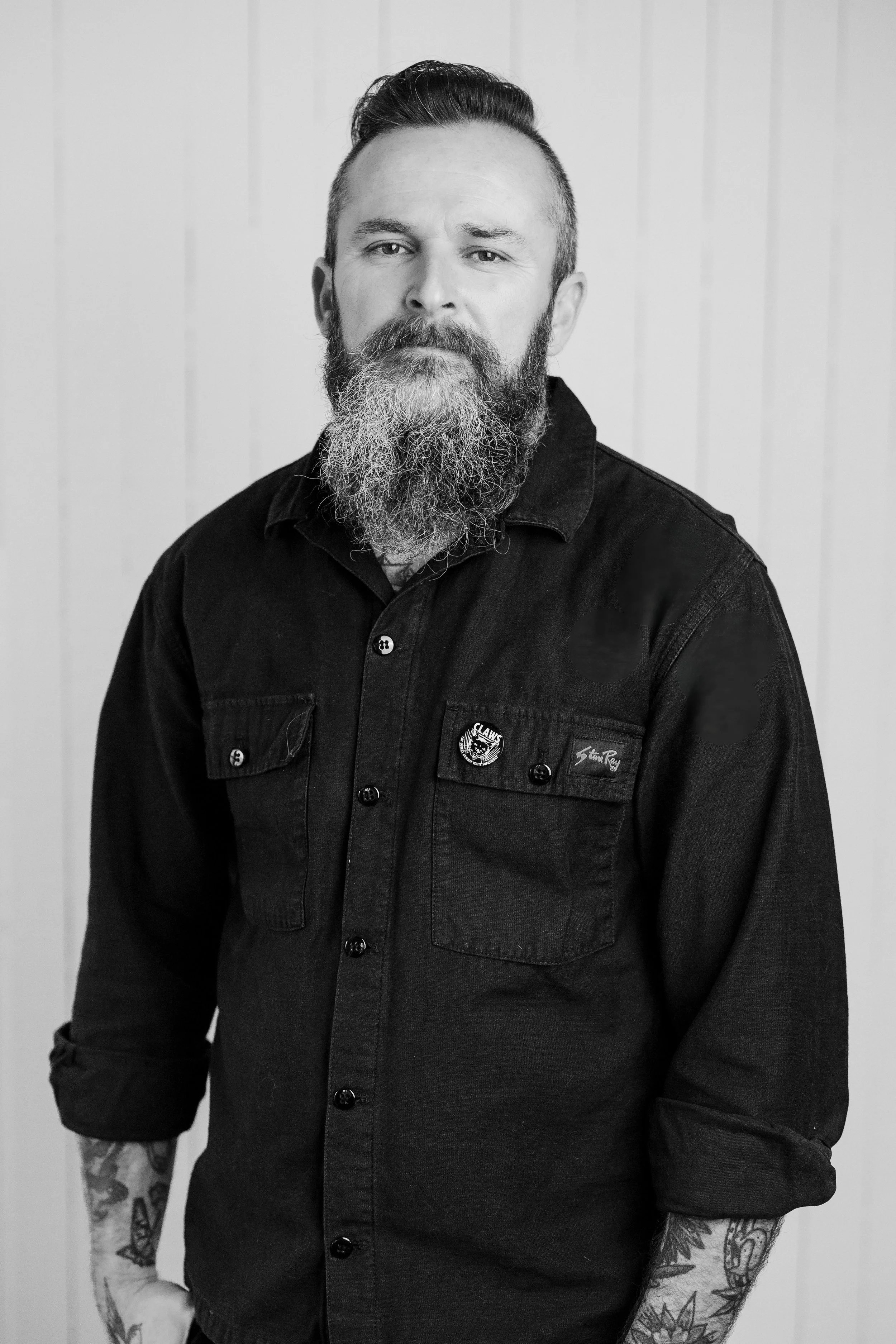 Black and white photo of a man with a beard and striped shirt, arms crossed, standing in front of a textured wall.