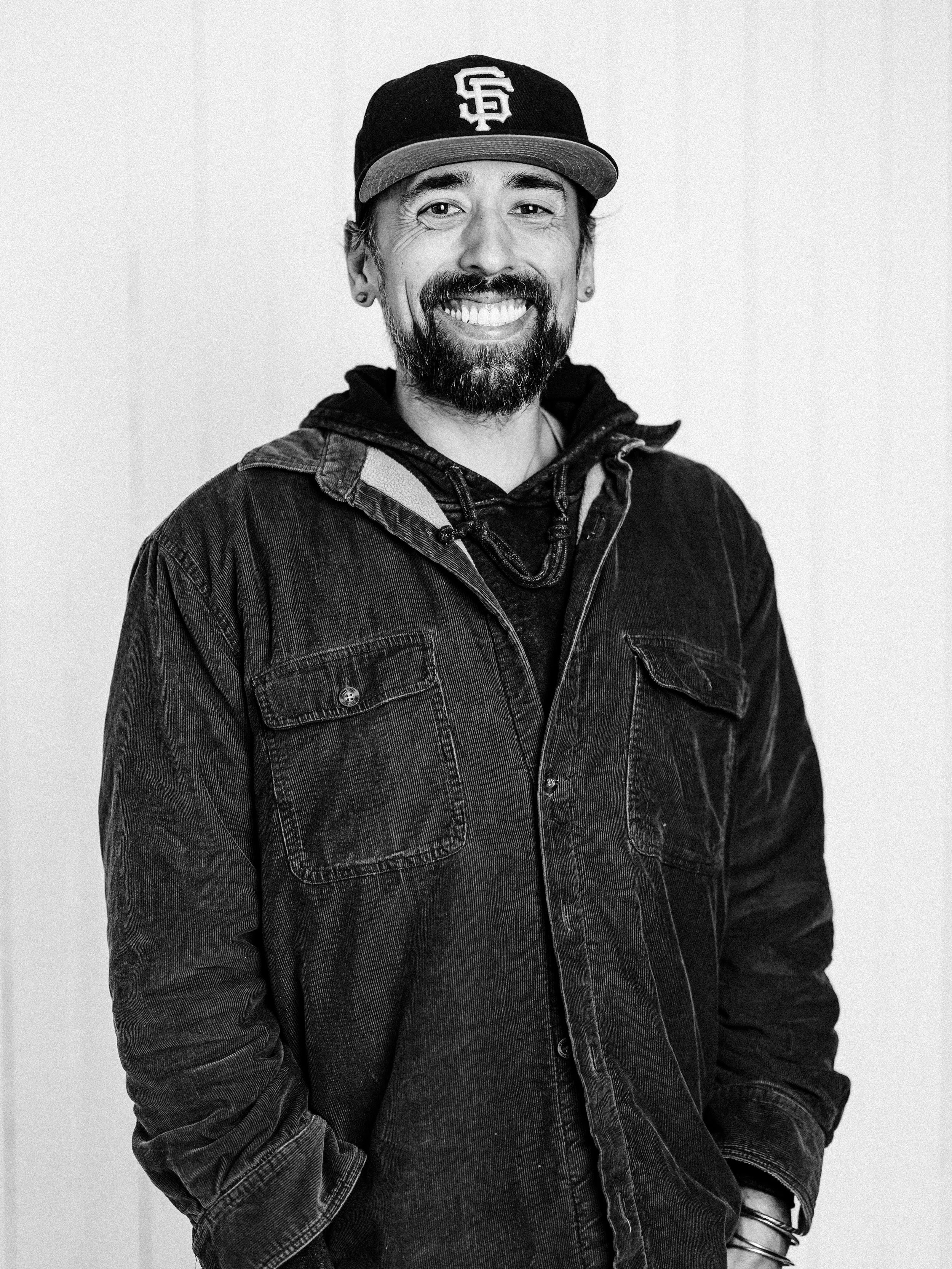 Black and white photo of a smiling man wearing a baseball cap and button-up shirt, standing in front of a wooden wall.
