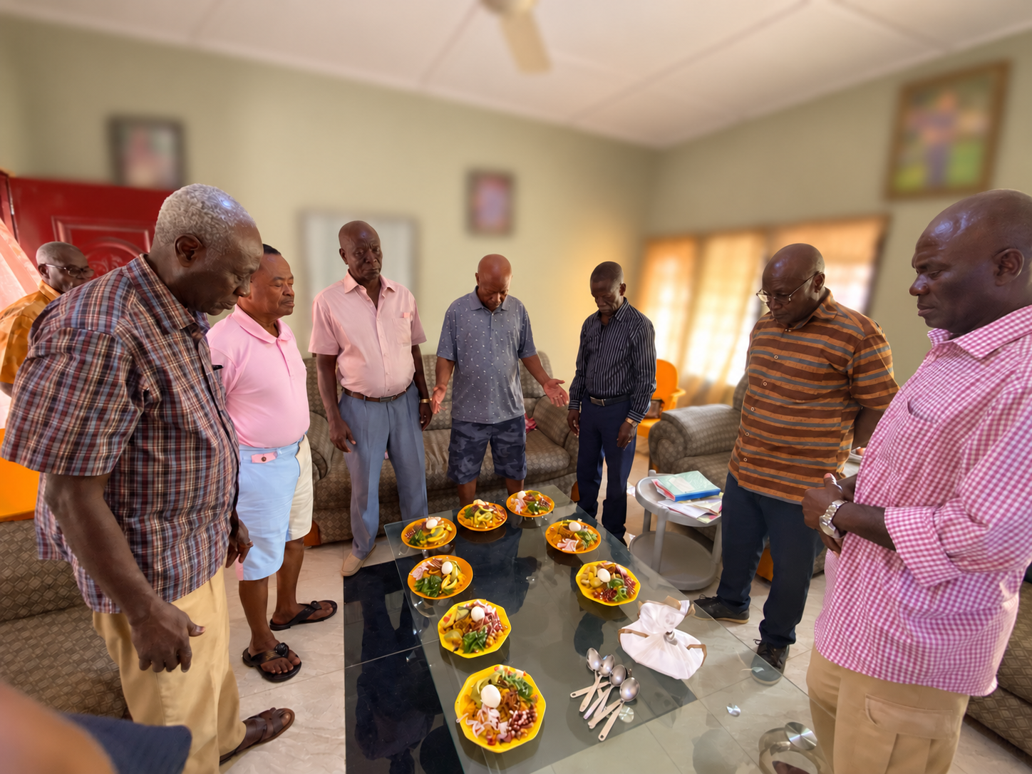 Men gathered around a glass table set with plates of food, several with their heads bowed in prayer.
