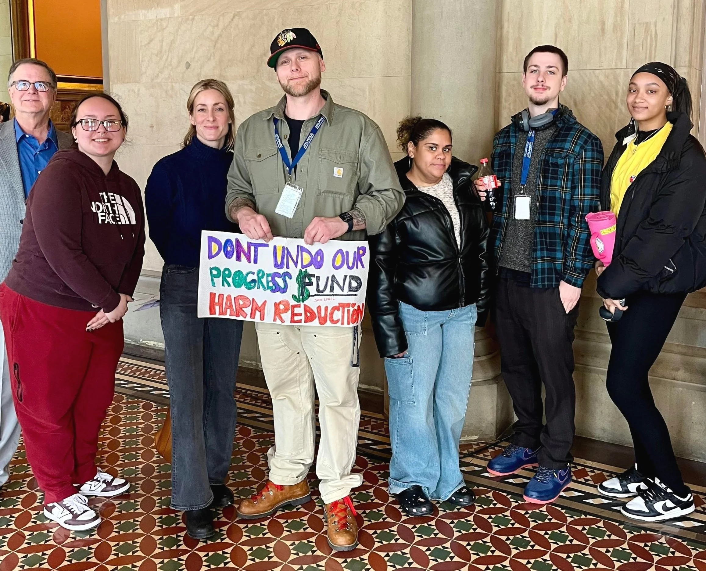 Group of seven people posing indoors with a sign reading “Don’t undo our progress, fund harm reduction.”