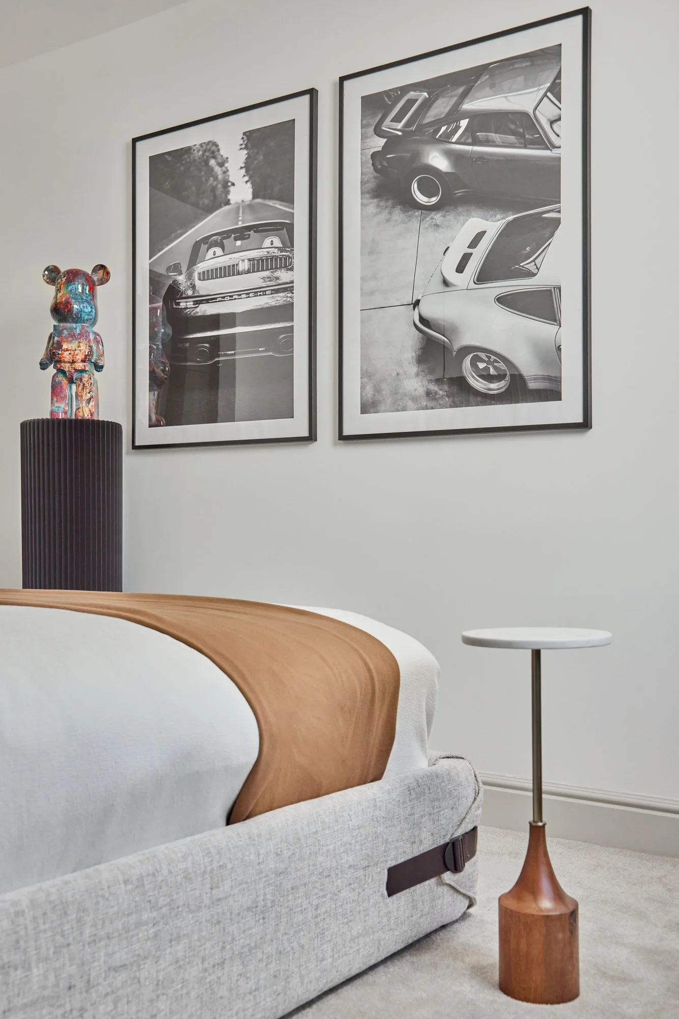 Modern bedroom interior in Wimbledon featuring a bed, artwork of vintage sports cars, a colourful bear figurine on a black pedestal, and a round side table with wooden base. A contemporary and playful space combining style with personality.