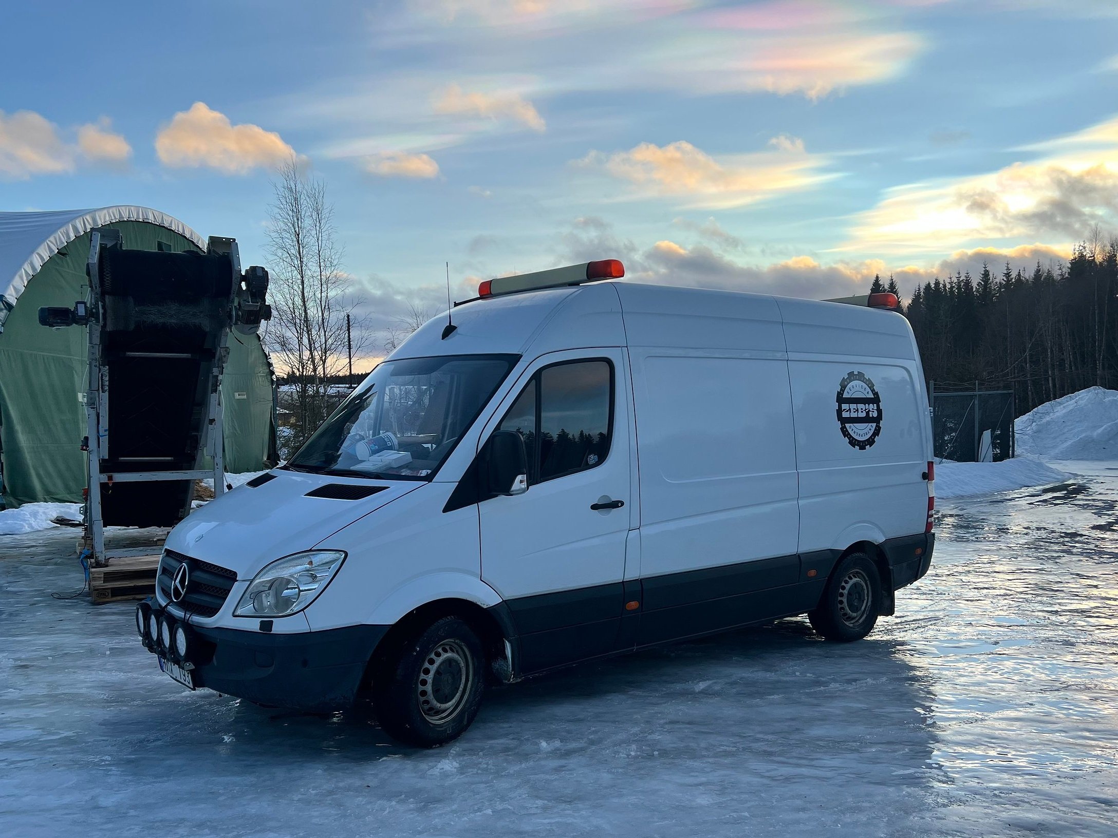 En vit Mercedes transportbil med logo från Zeb's Service och Entreprenad, parkering på isbelagd mark vid solnedgång, omgiven av snö och vinterlandskap.