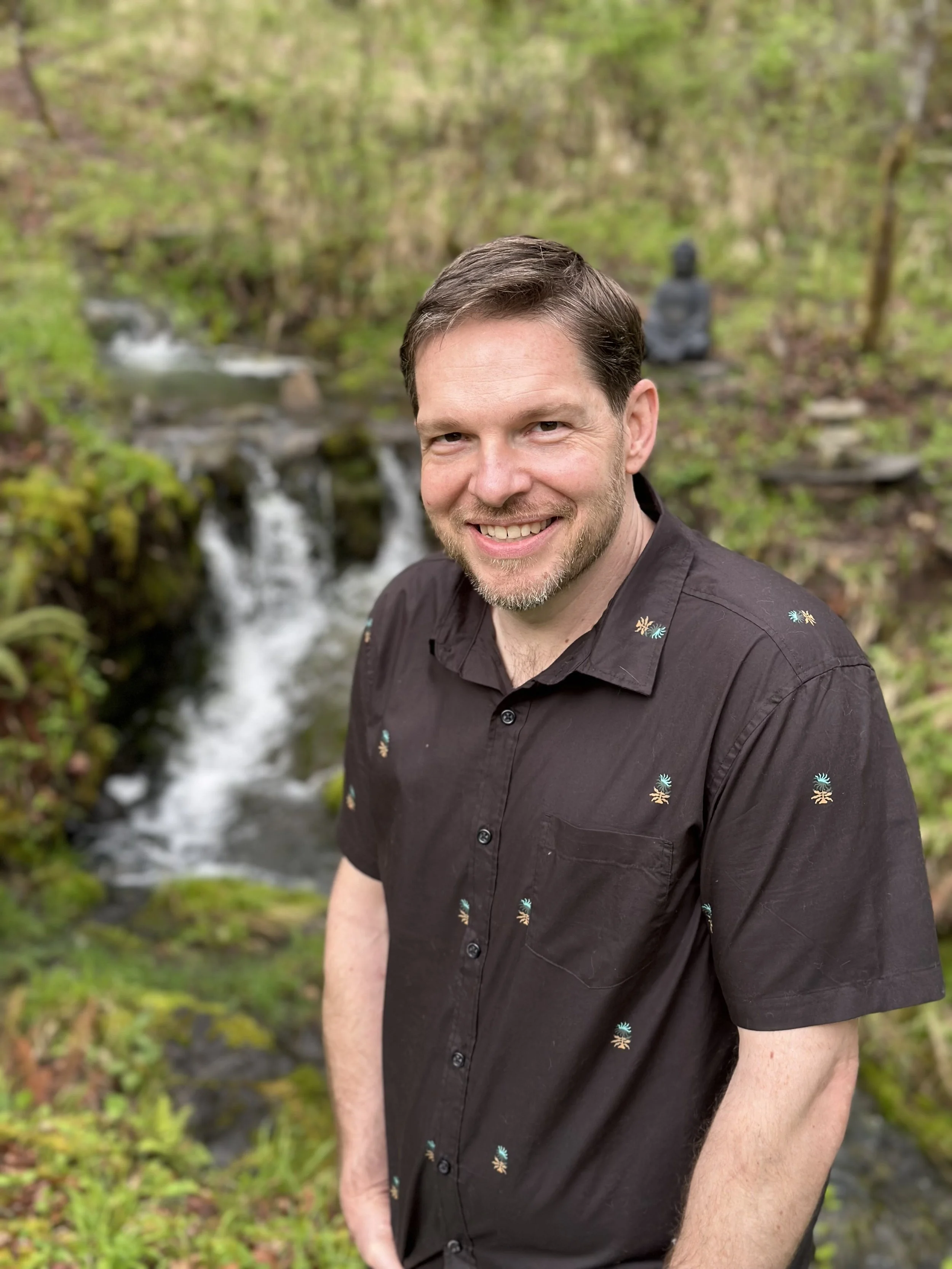 John, a man, in a dark shirt, smiling in front of a small waterfall and  foliage, with a buddha stone statue in the background.