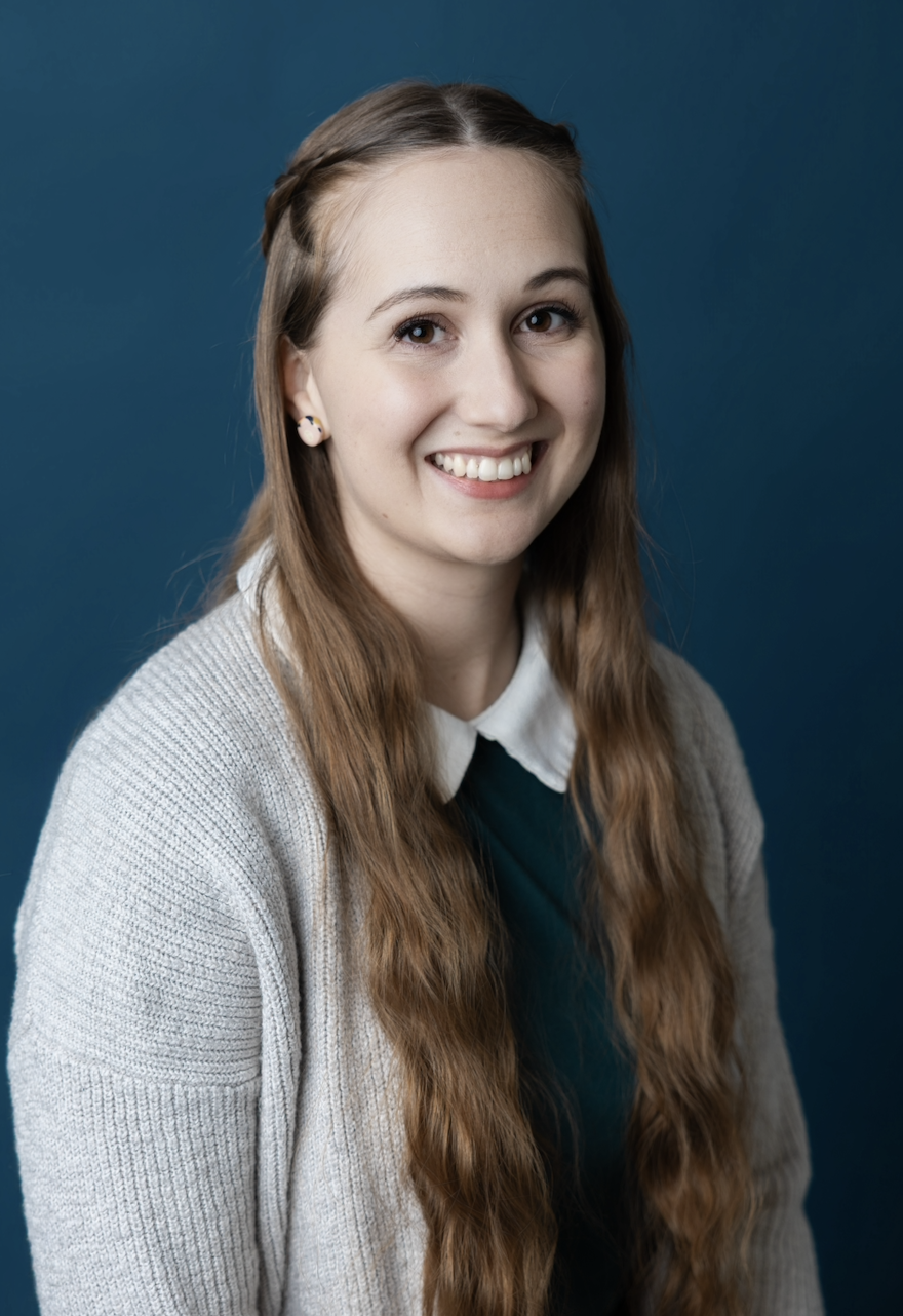 Portrait of a young woman with long, wavy brown hair, smiling, against a solid blue background, wearing a white collared shirt, black sweater, and beige cardigan.