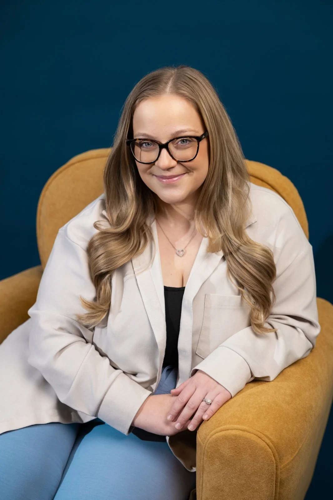 Smiling woman with long blond hair and glasses, sitting in a mustard yellow chair.