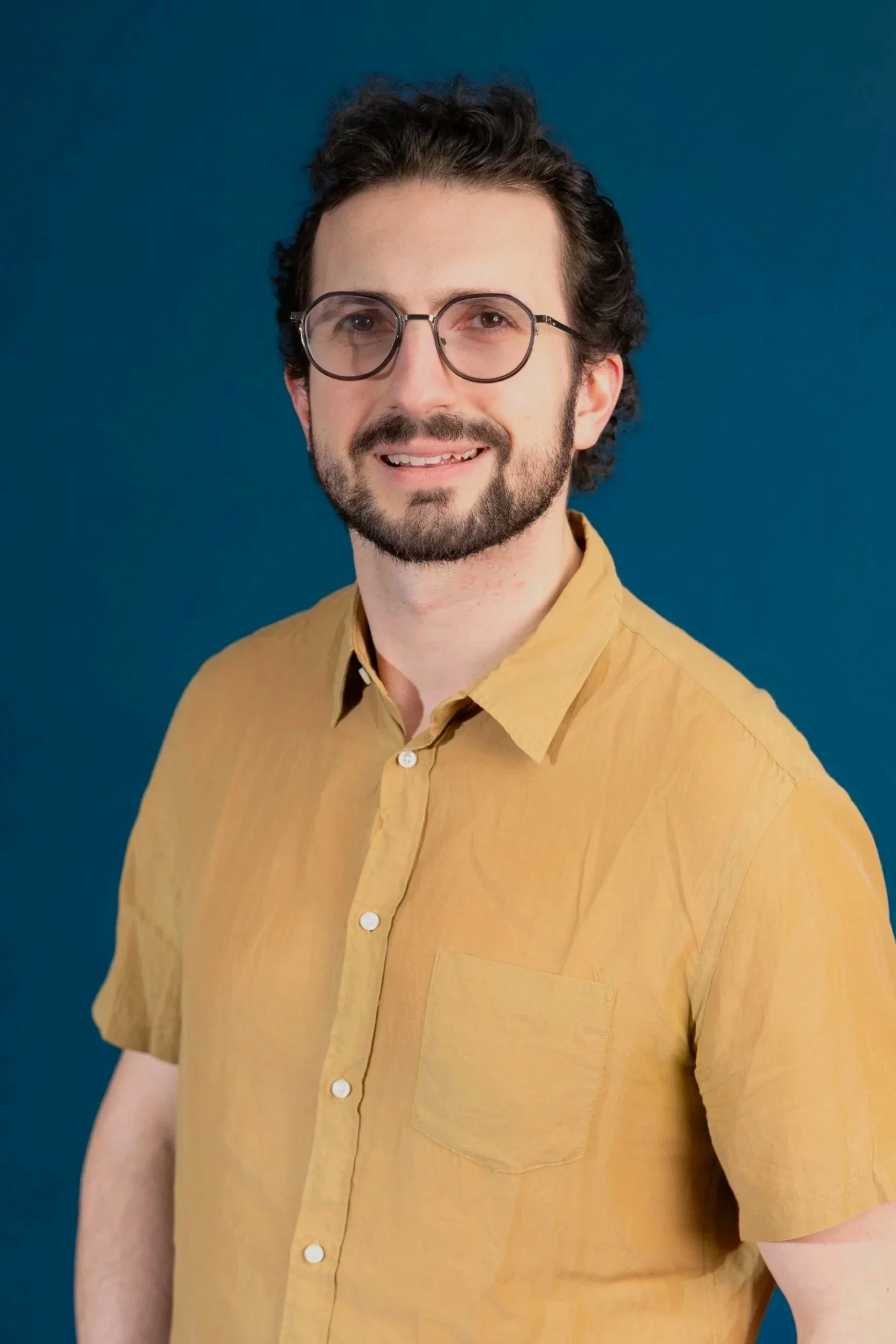 Man with brown hair and beard, smiling and wearing glasses.