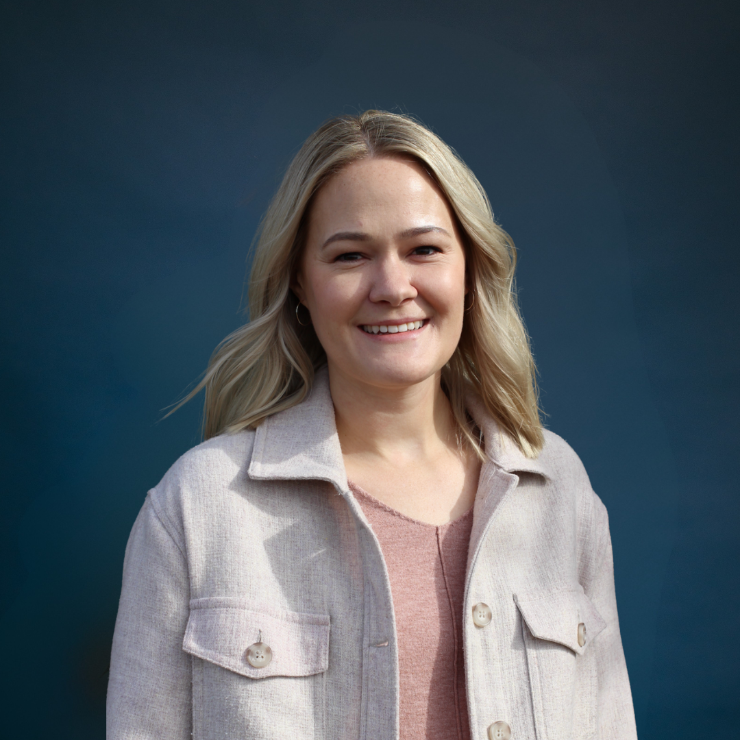 Smiling person outdoors wearing a light jacket, with trees in the background.