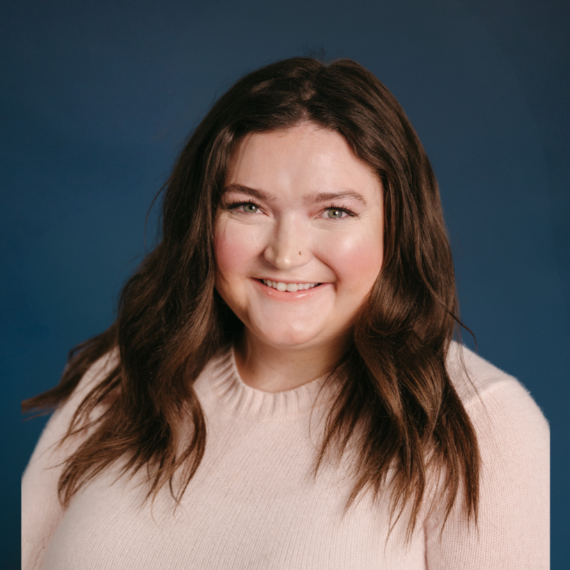 Woman smiling with long brown hair, wearing a light-colored sweater with heart patterns, leaning on a chair, against a blue background.