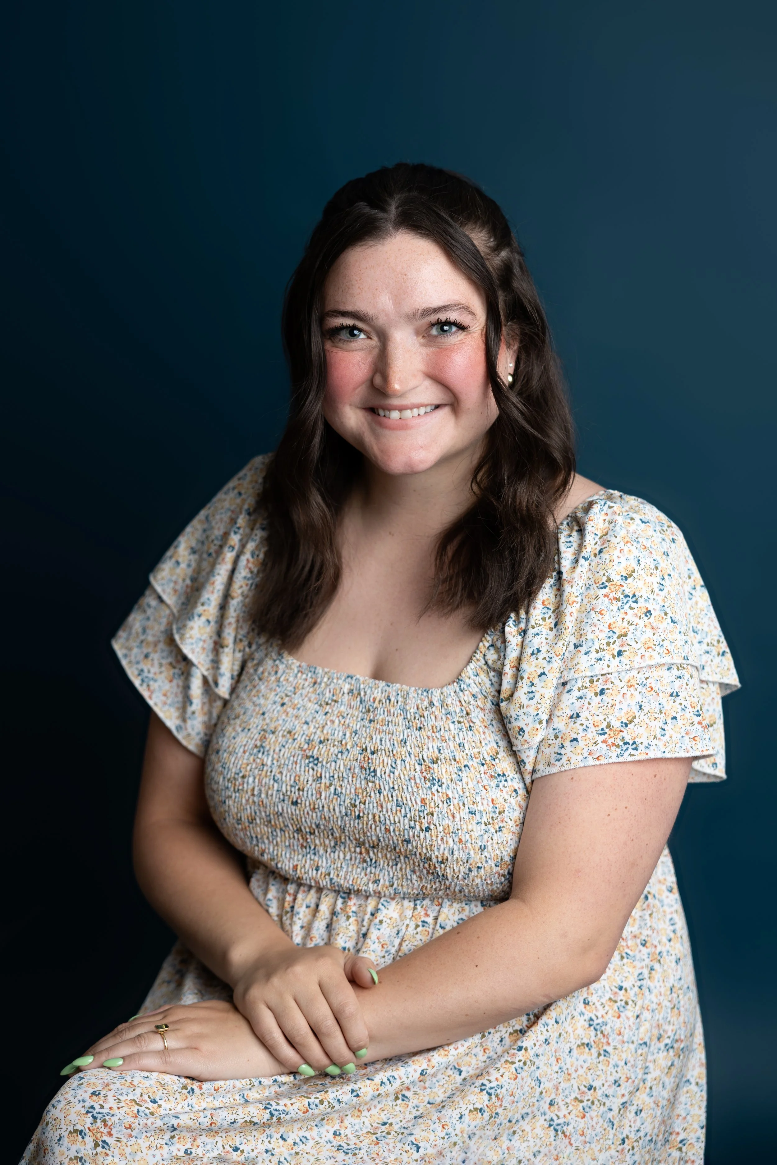 Woman smiling with long brown hair, wearing a light-colored sweater with heart patterns, leaning on a chair, against a blue background.