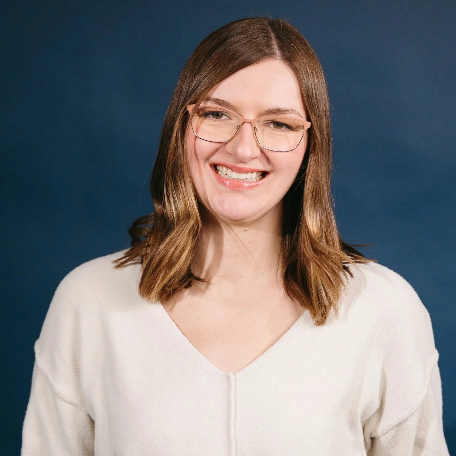 Edmonton Occupational Therapist with glasses and shoulder-length hair, wearing a white sweater and black pants, standing confidently against a dark blue background, specializing in sensory integration and motor skills therapy.