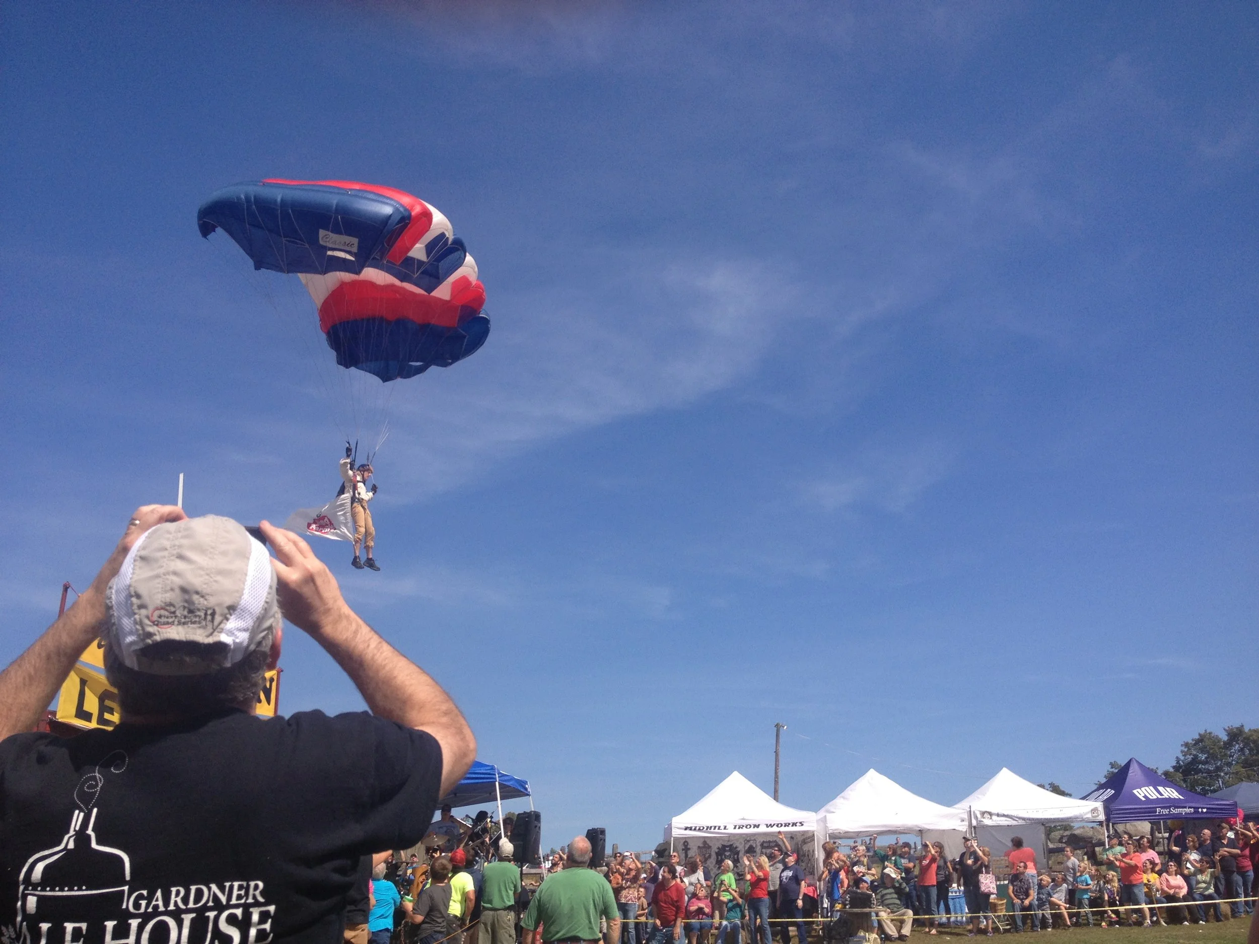 A parasailer dressed as Johnny Appleseed with a Red Apple Farm flag flying in a blue sky during a crowded outdoor event. Many people are gathered on the ground, some taking pictures and watching the parasailer.