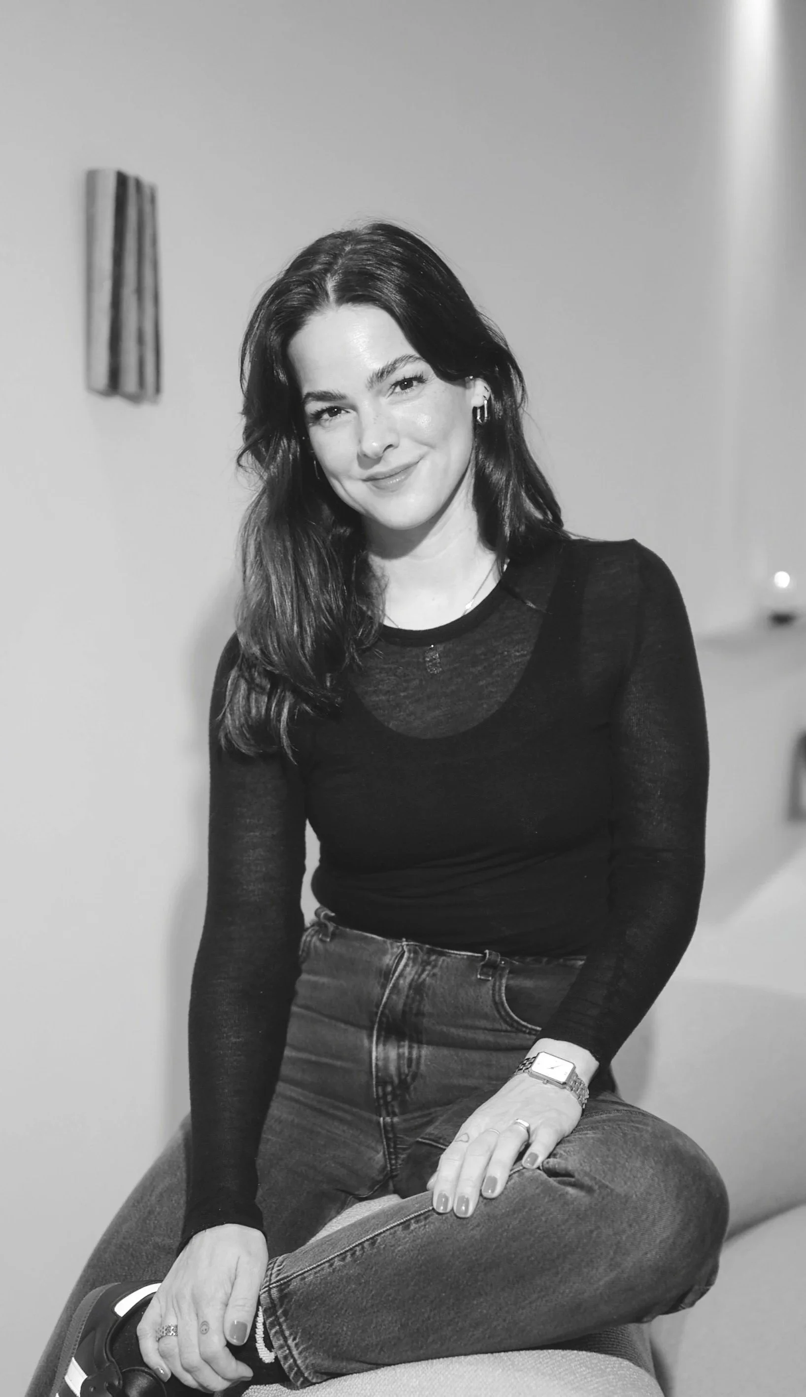 A woman with dark, shoulder-length hair, wearing a black long-sleeve top and jeans, sitting cross-legged on a piece of furniture, smiling at the camera in a monochrome photo.
