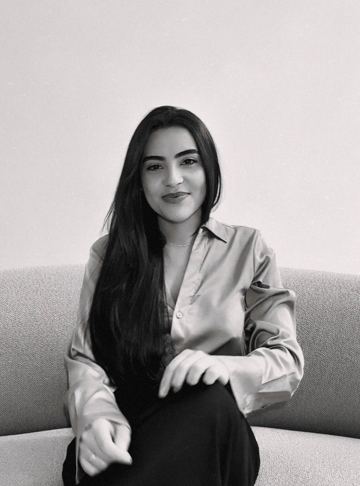 A woman with long dark hair, wearing a satin shirt, sitting on a light-colored couch, smiling at the camera in a black-and-white photo.