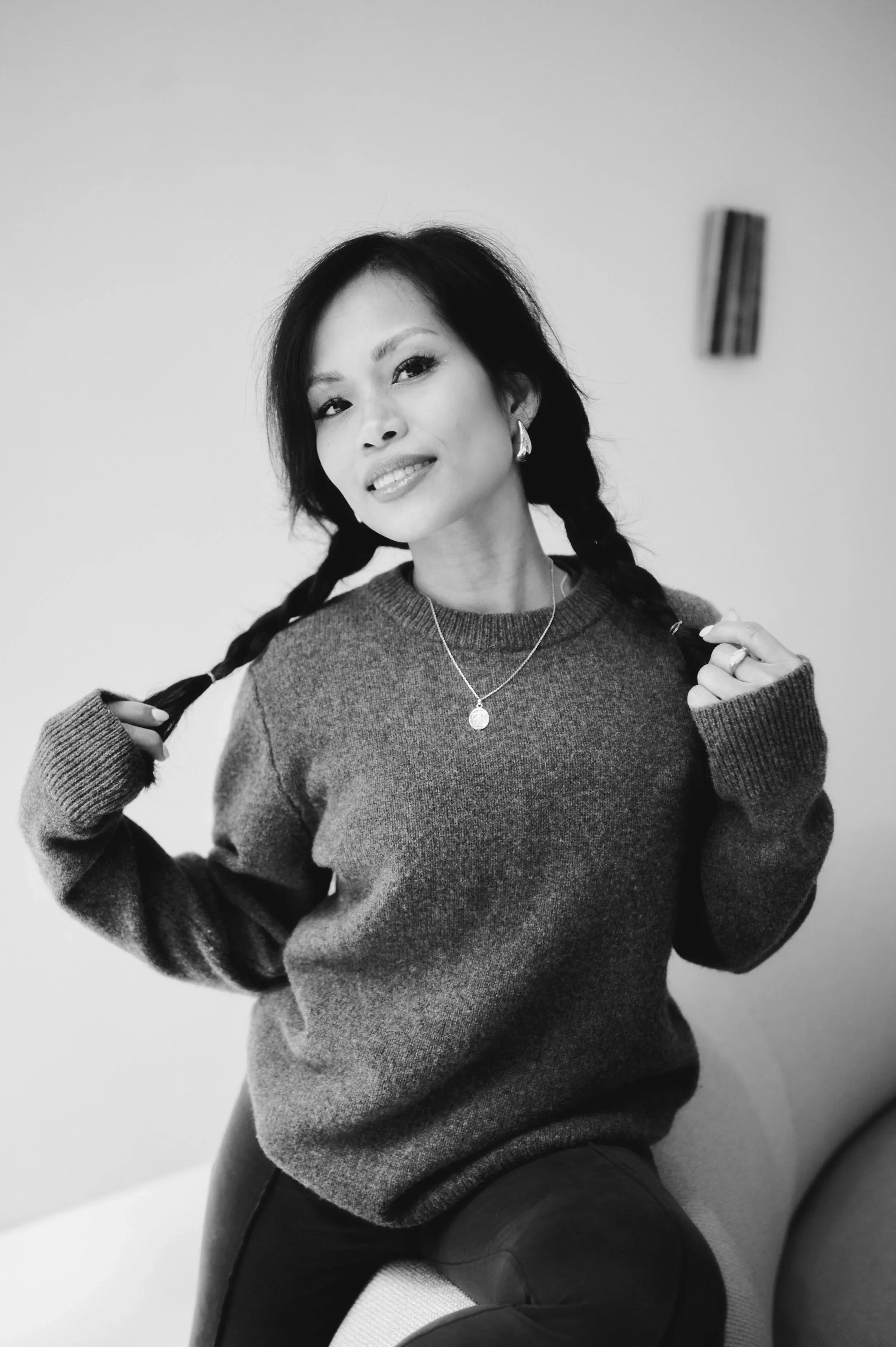 A woman with dark hair in braids wearing a sweater and jewelry, sitting on a chair against a plain background.
