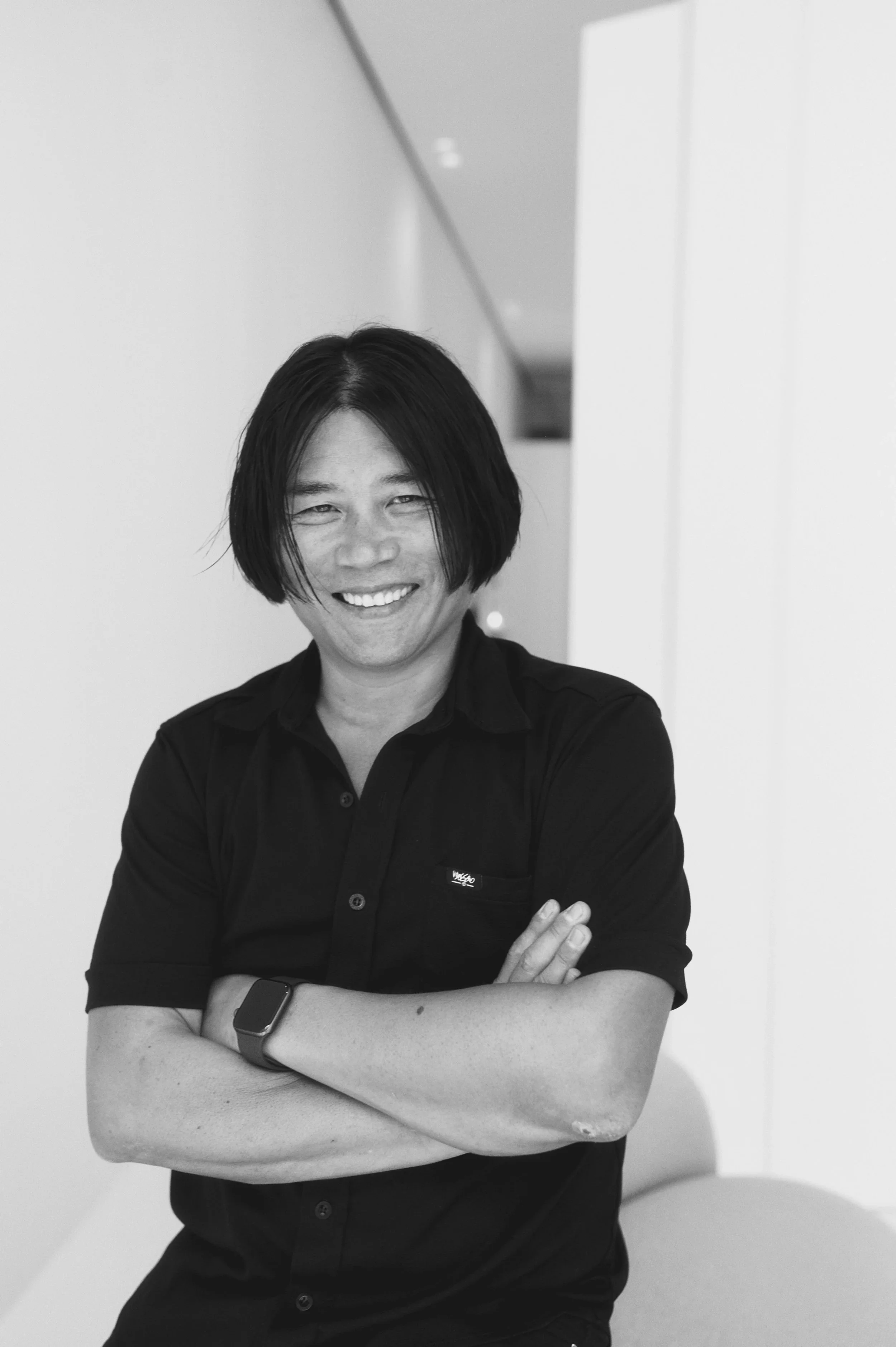 Black and white photo of a smiling man with medium-length hair, wearing a black shirt with his arms crossed, standing in a modern, minimalist indoor space.