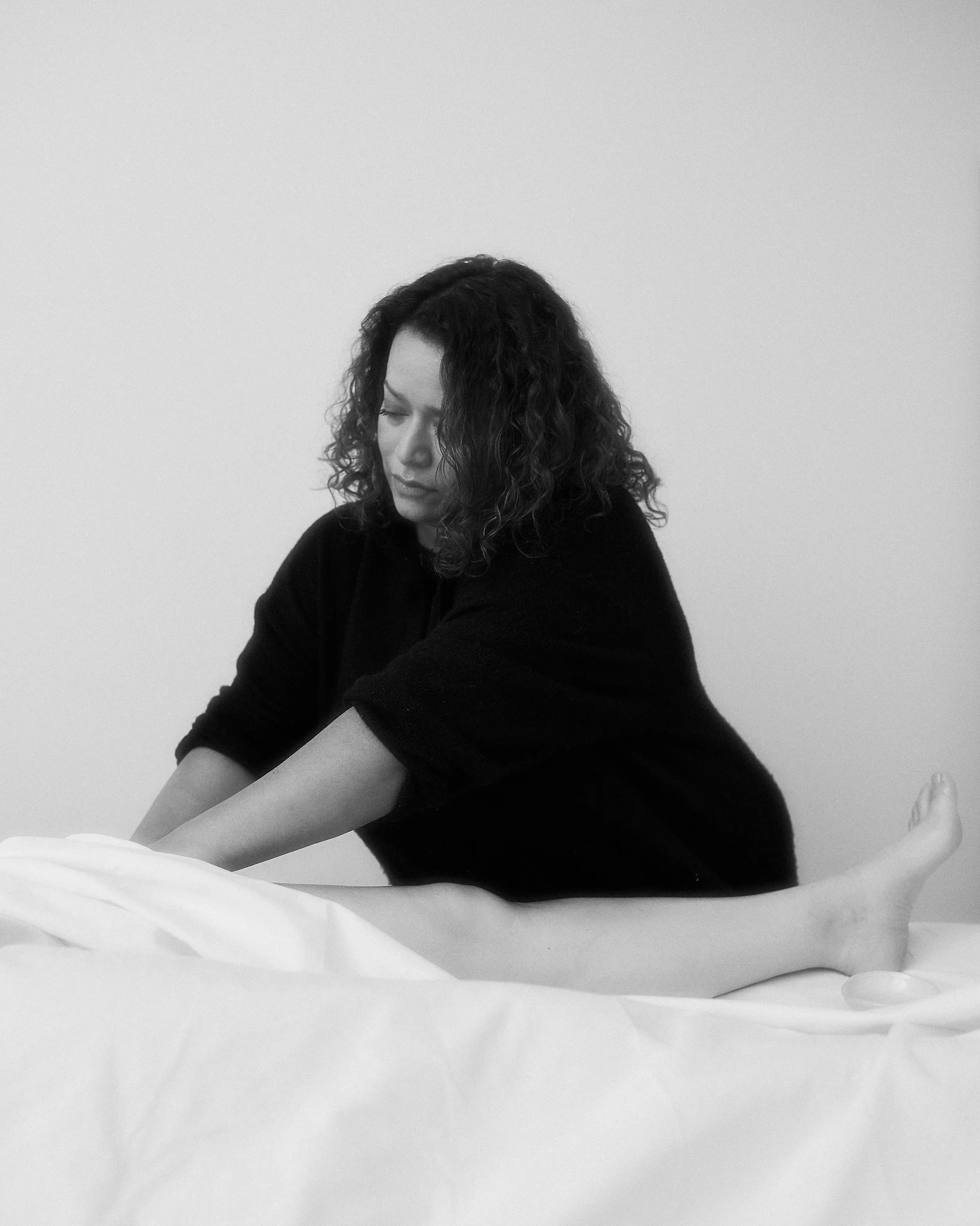 A woman with curly hair sitting on a bed, stretching her legs while looking down, in black and white.