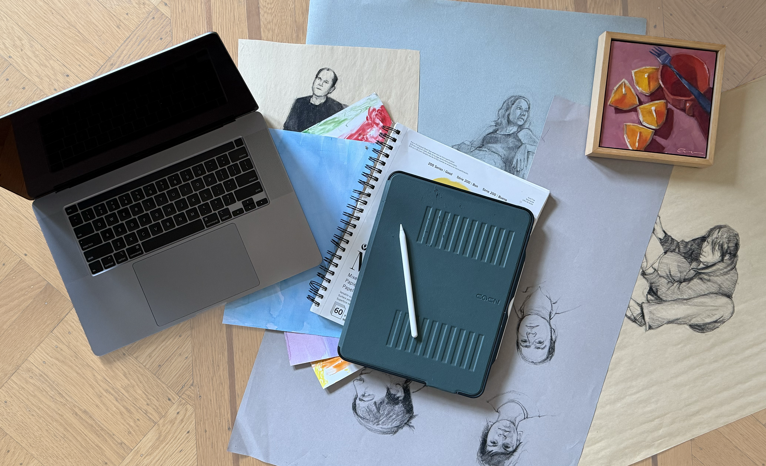 Overhead view of a laptop, sketches, art supplies, and a framed painting on a wooden floor. The laptop is open beside several sketch papers with pencil drawings of human figures. An iPad with a stylus rests on a drawing pad. A colorful framed artwork depicting orange slices is also visible.