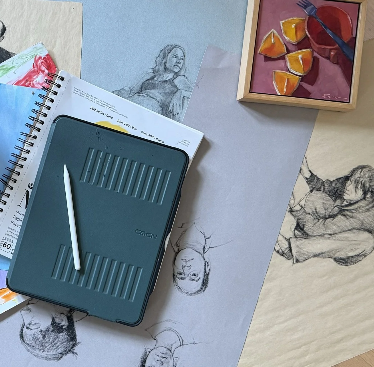 Artistic workspace with sketches, sketchpad, digital tablet, and framed painting. Various figure drawings on paper are spread out, accompanied by a stylus on the tablet and mixed media sketchpad.