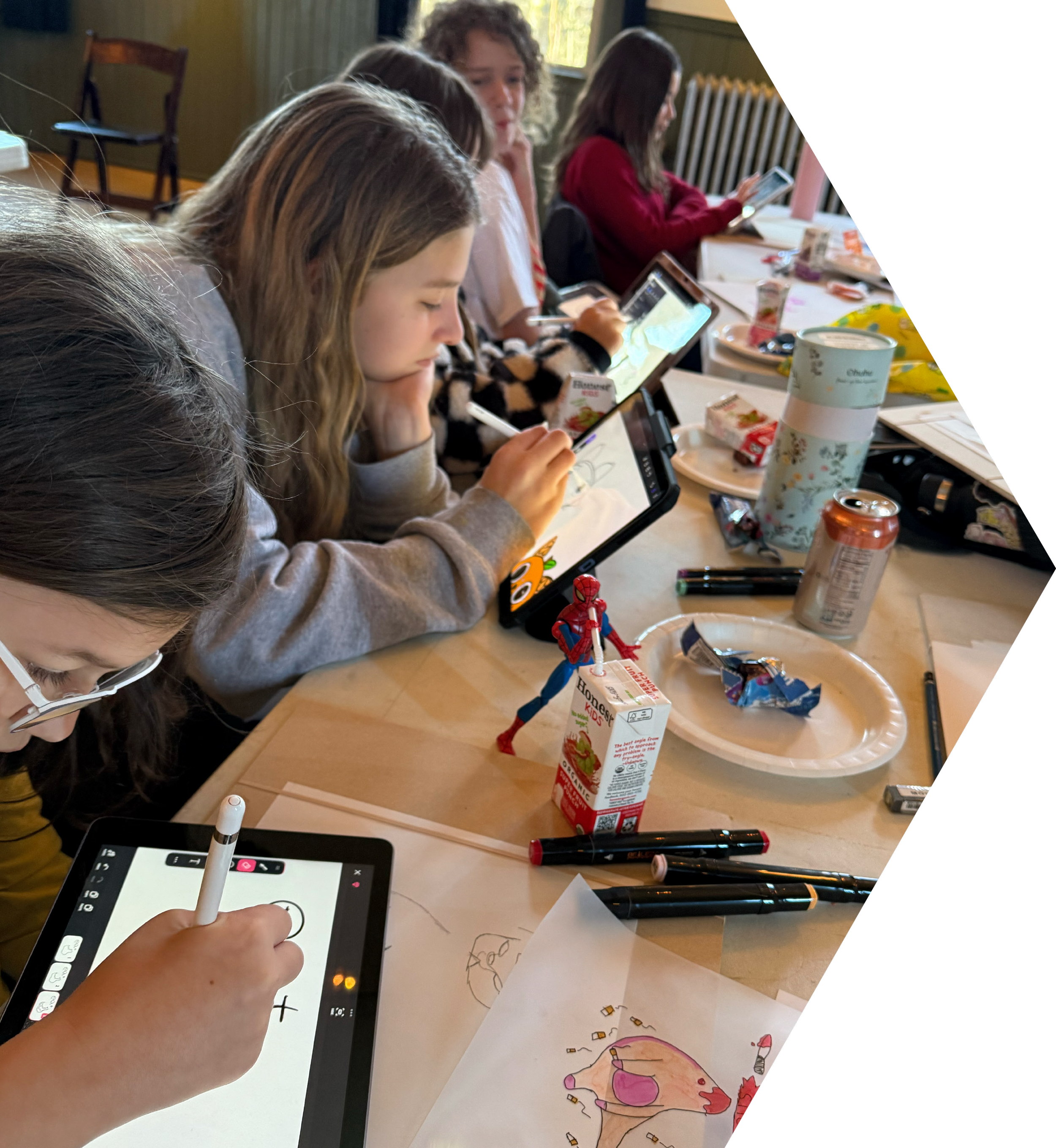 Children drawing on tablets with styluses at a table with snacks and drinks.