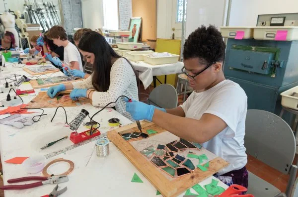 A group of people working on stained glass projects at a table, using soldering tools and wearing blue gloves.