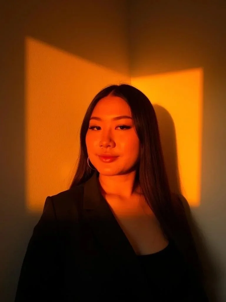 A woman with long dark hair and hoop earrings taking a selfie against a wall with orange and yellow sunset lighting patterns.
