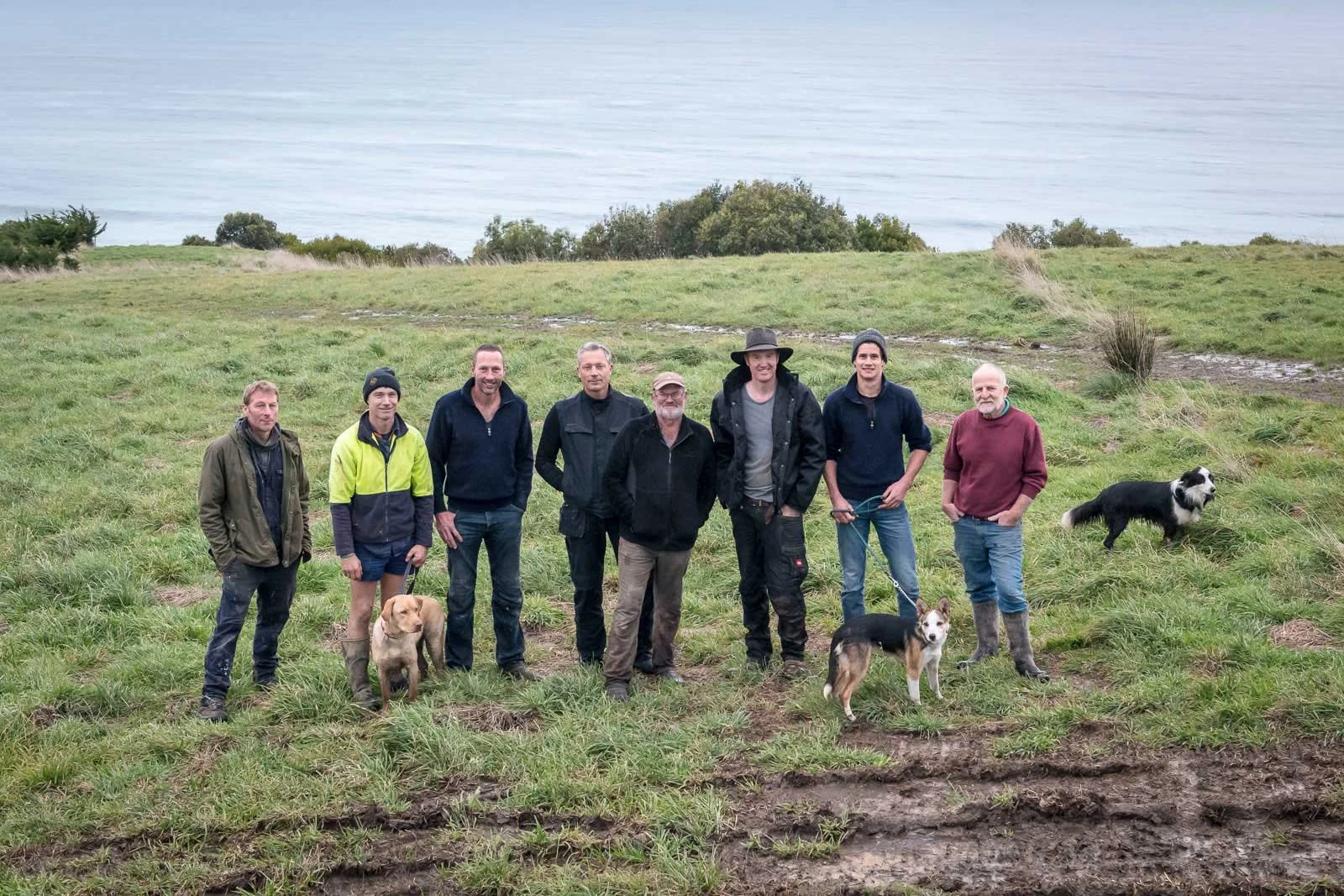 Eight men standing on a grassy field by the ocean, with three dogs. They are dressed in casual outdoor clothing.