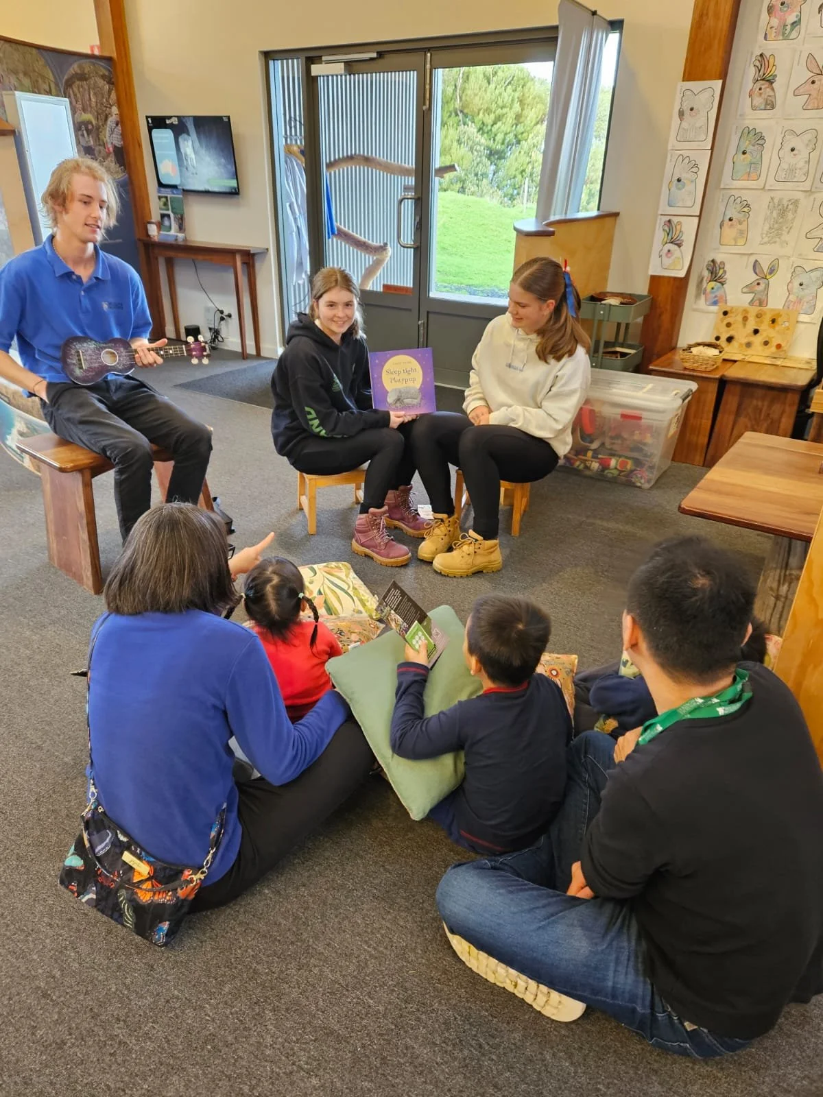 Group of adults and children gathered in a room for a storytelling session; one person holds a book, another holds a ukulele, with colorful drawings on the wall.