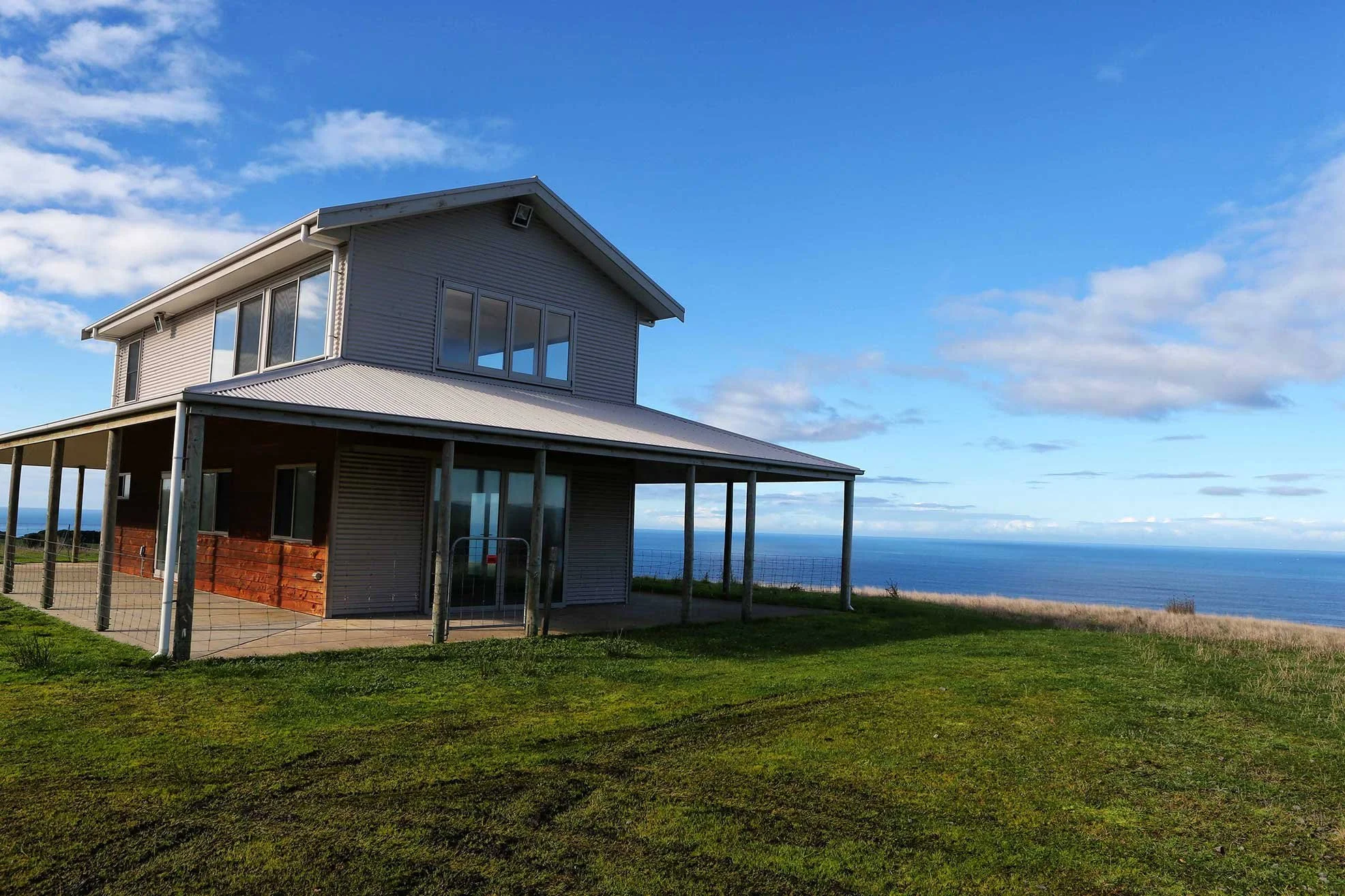 Two-story house with large windows overlooking the ocean, surrounded by green lawn, under a blue sky with clouds.